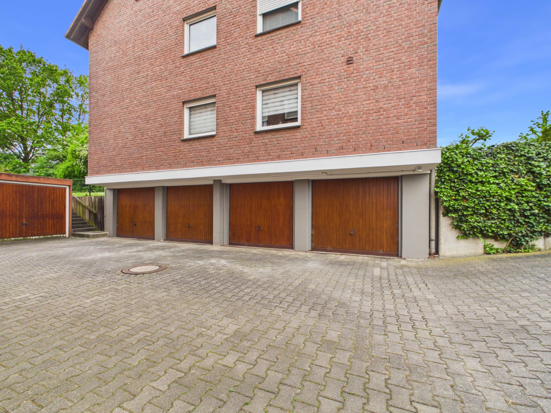 Mehrfamilienhaus mit vier Garagen und gepflastertem Hof, im Hintergrund Bäume und Begrenzungsmauer.