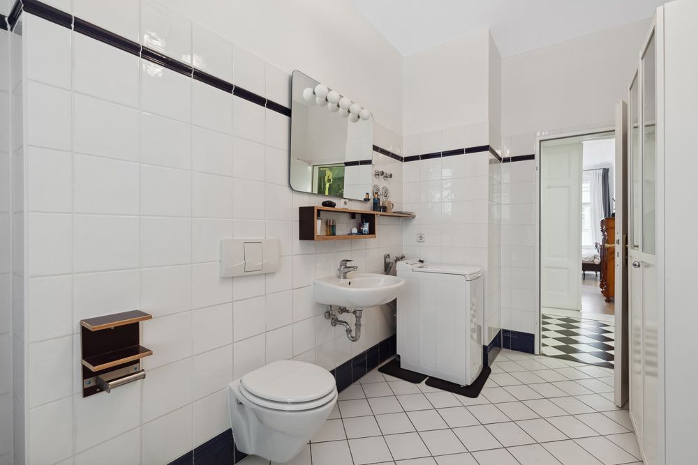 Helles Badezimmer mit WC, Waschbecken, Waschmaschine und Spiegel, weiße Fliesen an Wand und Boden.