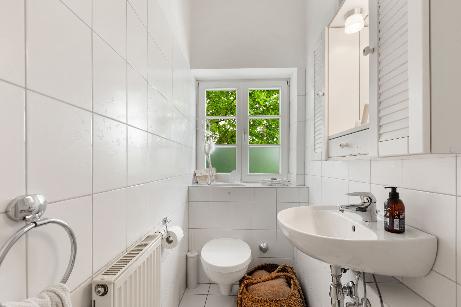 Helles, schmales Badezimmer mit weißer Fliesenwand, WC, Waschbecken und Fenster mit Blick ins Grüne.
