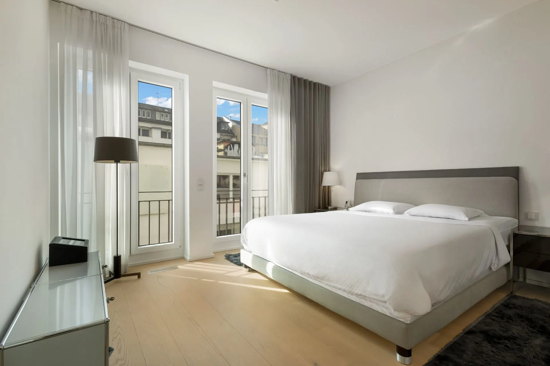 Modernes Schlafzimmer mit großem Bett, zwei Fenstern, Vorhängen und Nachttischen.