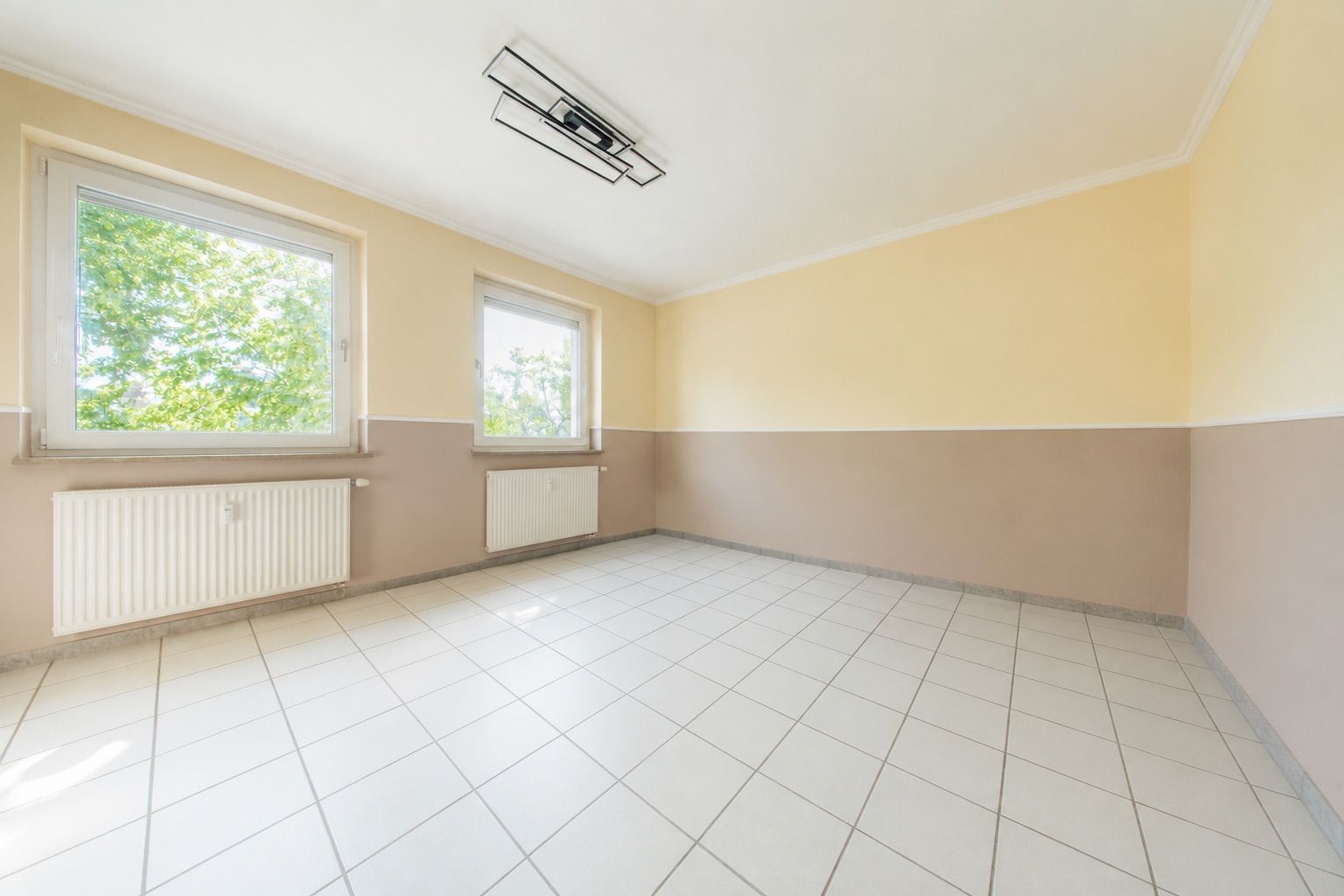 Leerer, heller Raum mit Fliesenboden, zwei Fenstern und Heizkörpern an der Wand.