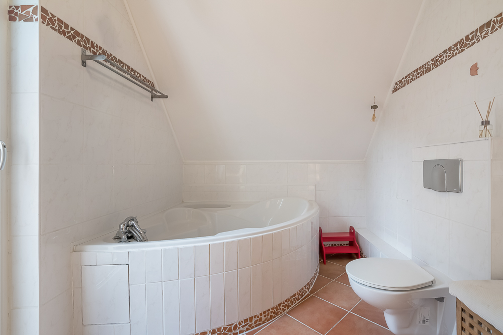 Helles Badezimmer mit Eckbadewanne, Toilette und rotem Tritthocker auf Fliesenboden.