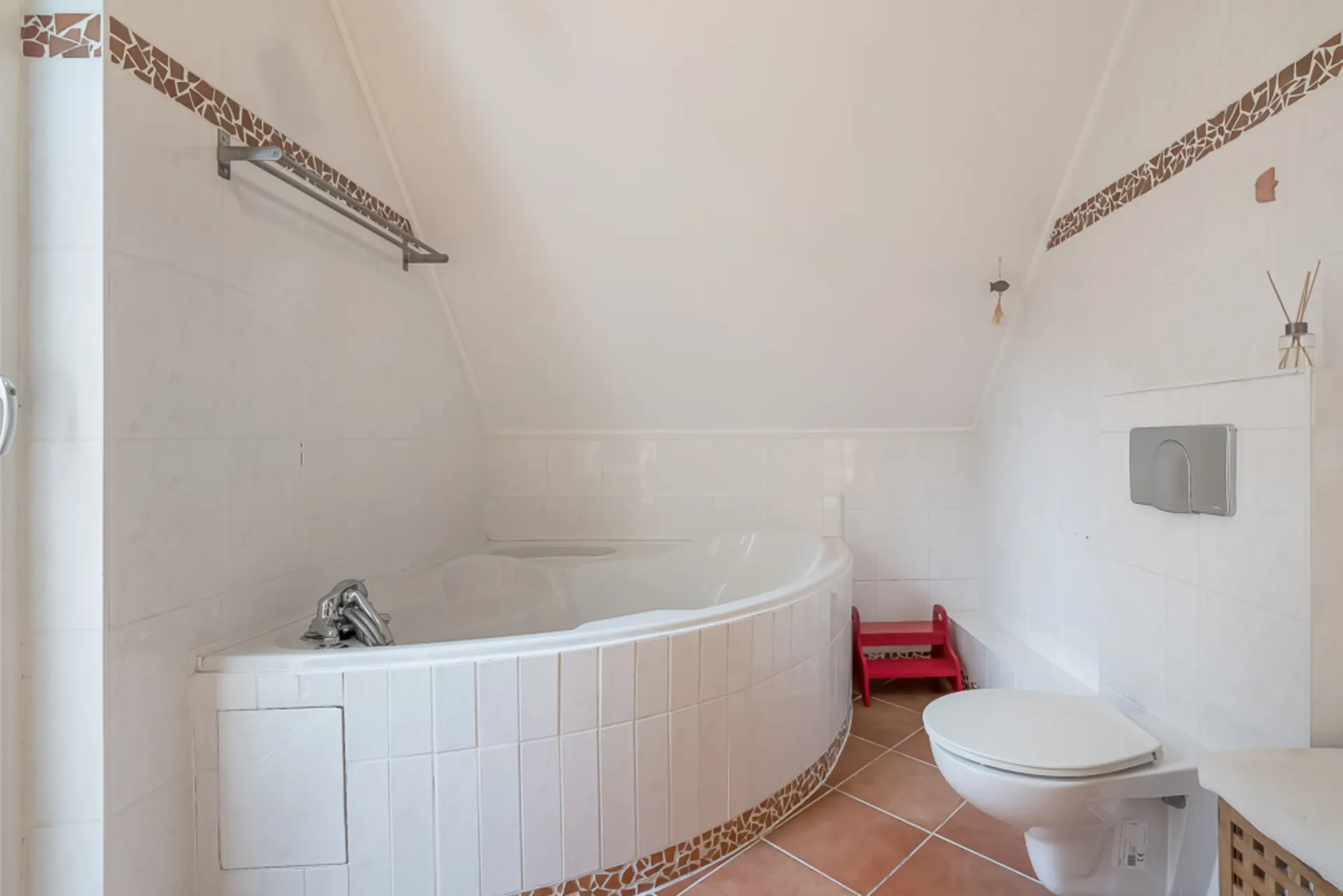 Helles Badezimmer mit Eckbadewanne, Toilette und rotem Tritthocker auf Fliesenboden.