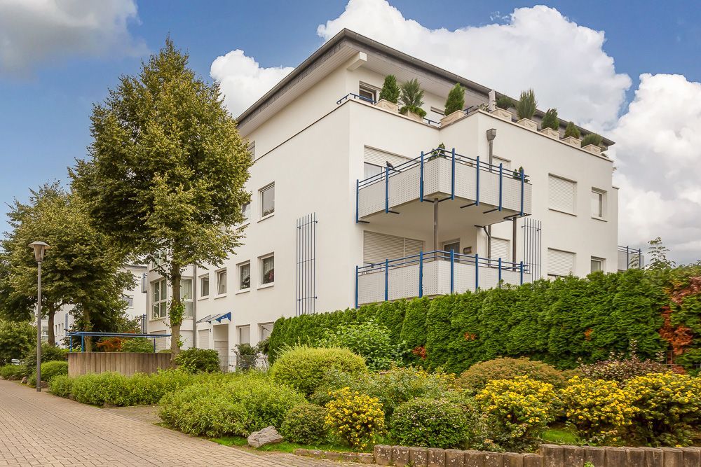 Modernes Mehrfamilienhaus mit mehreren Balkonen, umgeben von Bäumen und gepflegten Büschen.