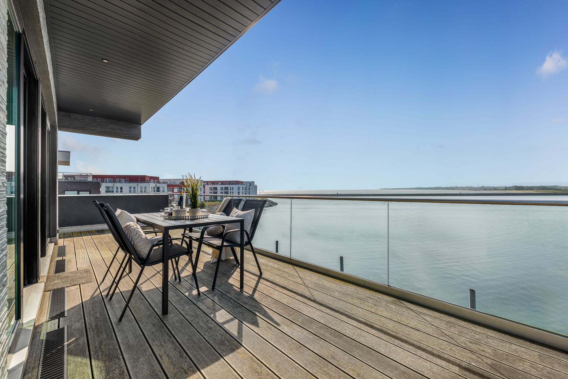 Großer Balkon mit Holzboden, Glengeländer und Esstisch mit vier Stühlen am Wasser.
