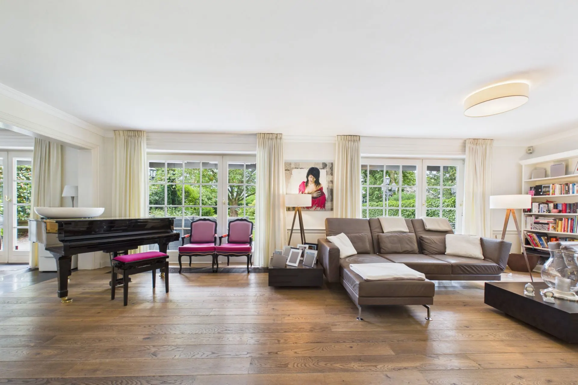 Helles Wohnzimmer mit Sofa, Flügel, Bücherregal und großen Fenstern mit Blick ins Grüne.