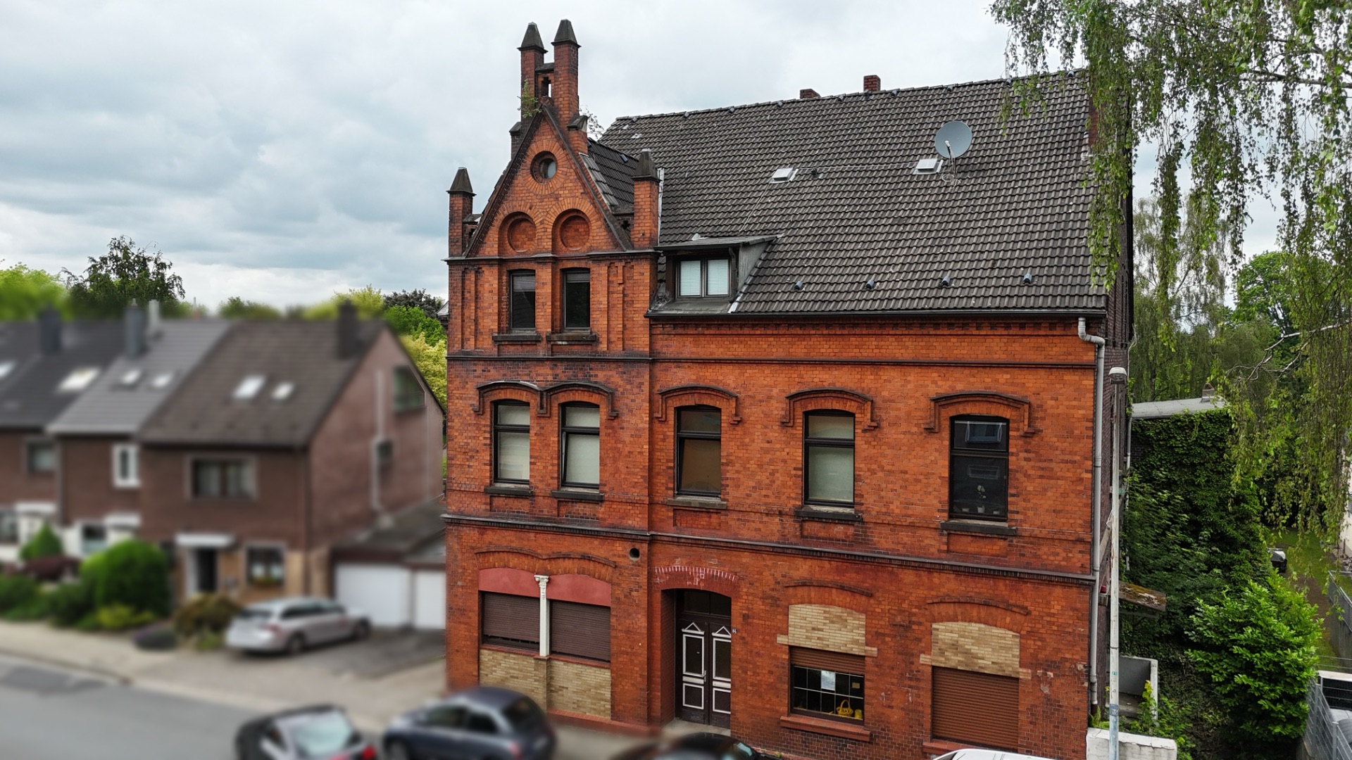 Rotes Backsteinhaus mit mehreren Etagen und verschlossenen Rollläden, daneben Wohnhäuser und Autos.