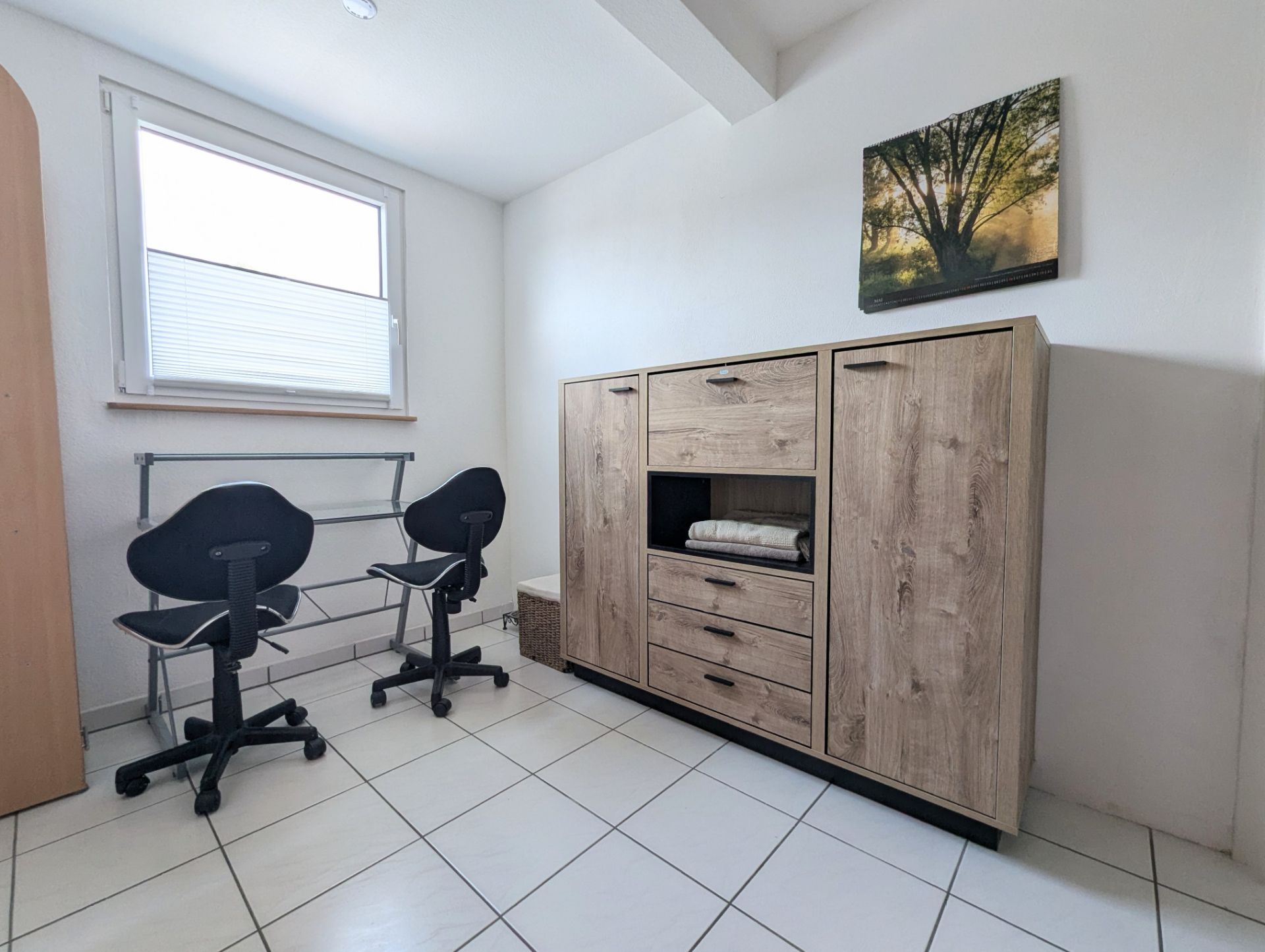 Heller Raum mit zwei schwarzen Stühlen, Holzkommode und Bild eines Baumes an der Wand.