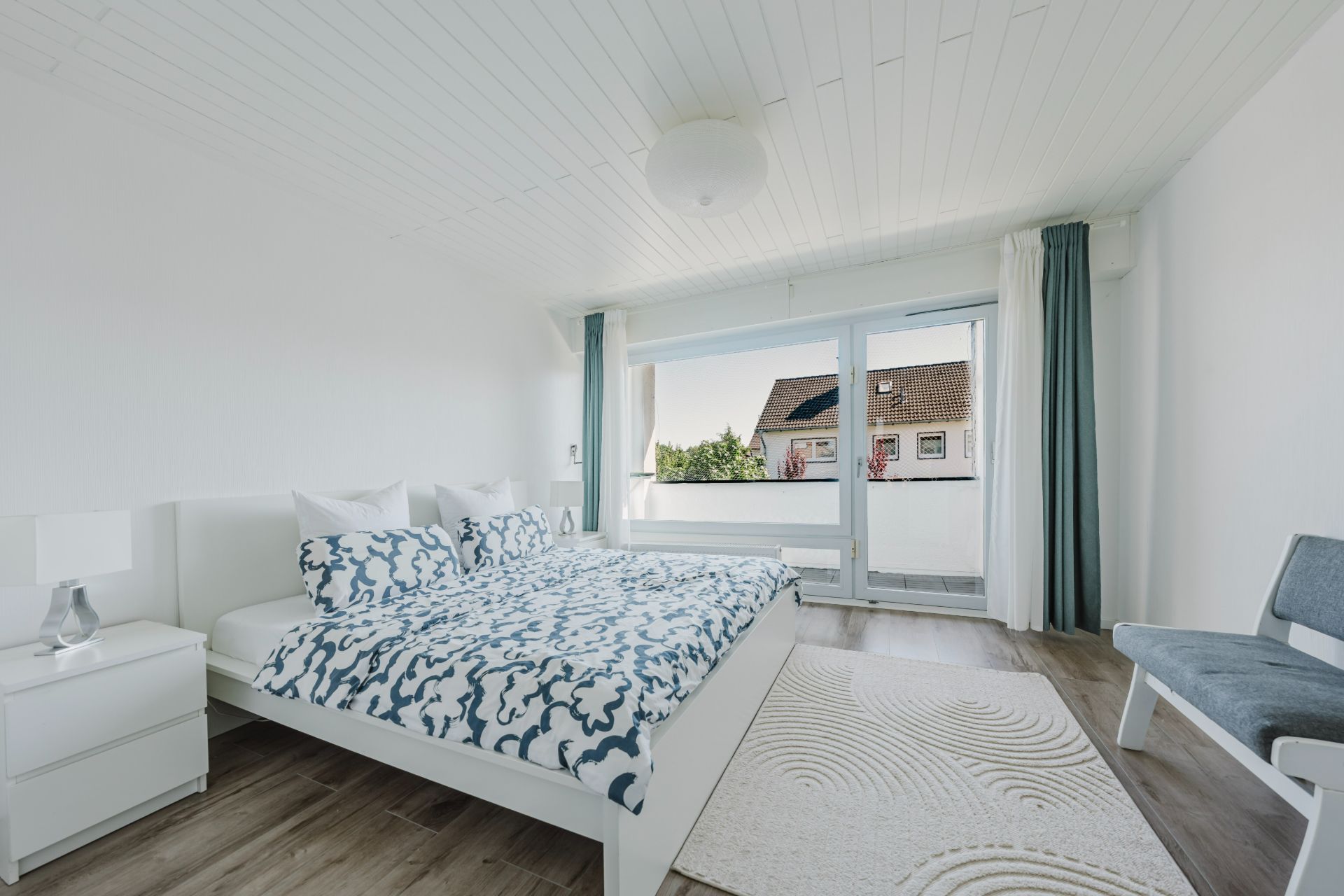 Modernes Schlafzimmer mit Doppelbett, großem Fenster und weißem Teppich auf Holzboden.