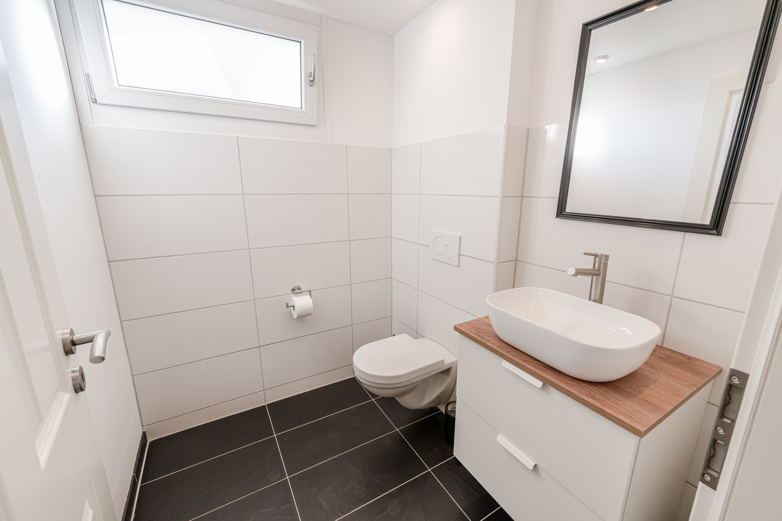 Modernes, weiß gefliestes Badezimmer mit Wandtoilette, Waschbecken und Spiegel.