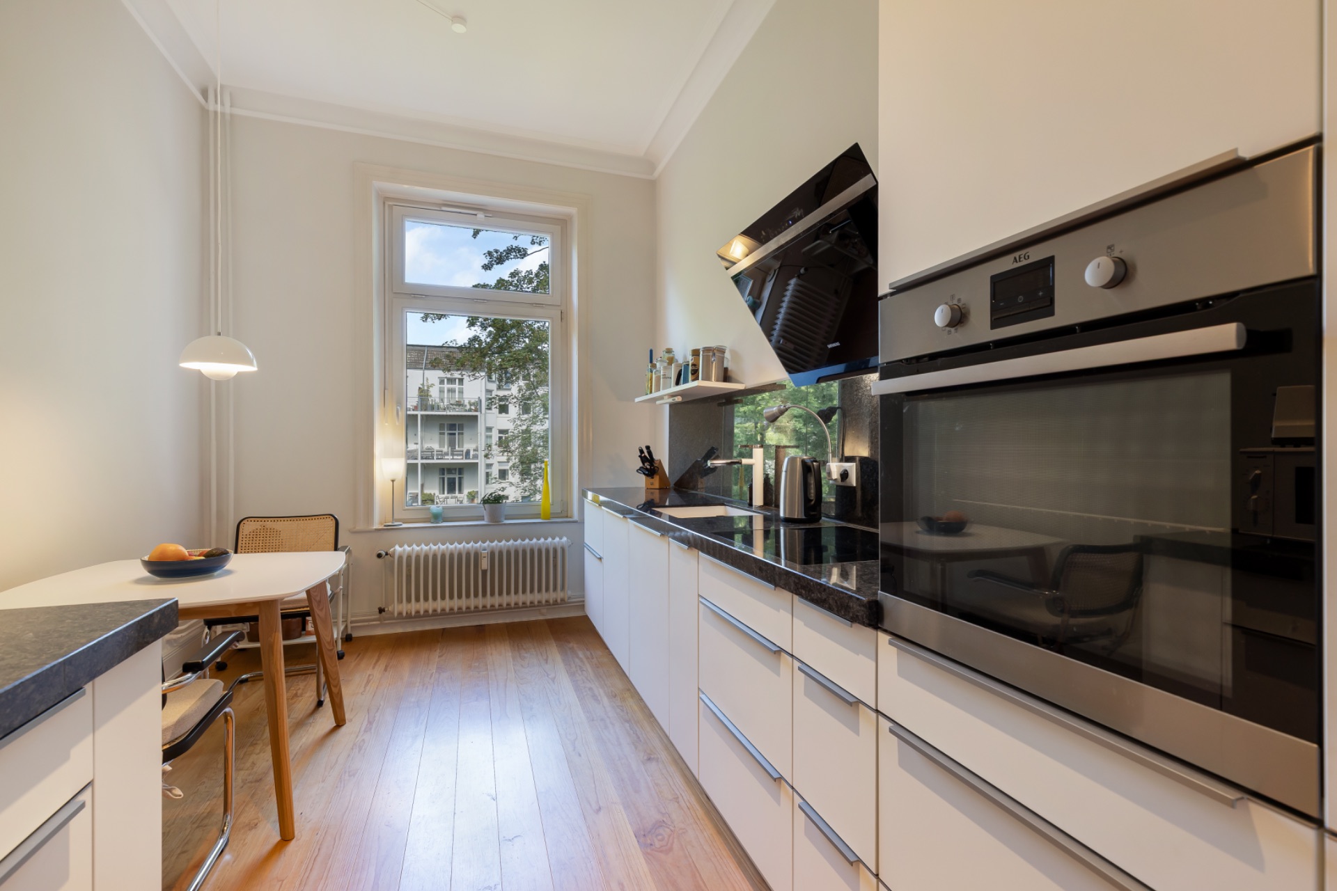 Moderne Küche mit weißen Schränken, Holzfußboden, Esstisch und großem Fenster.