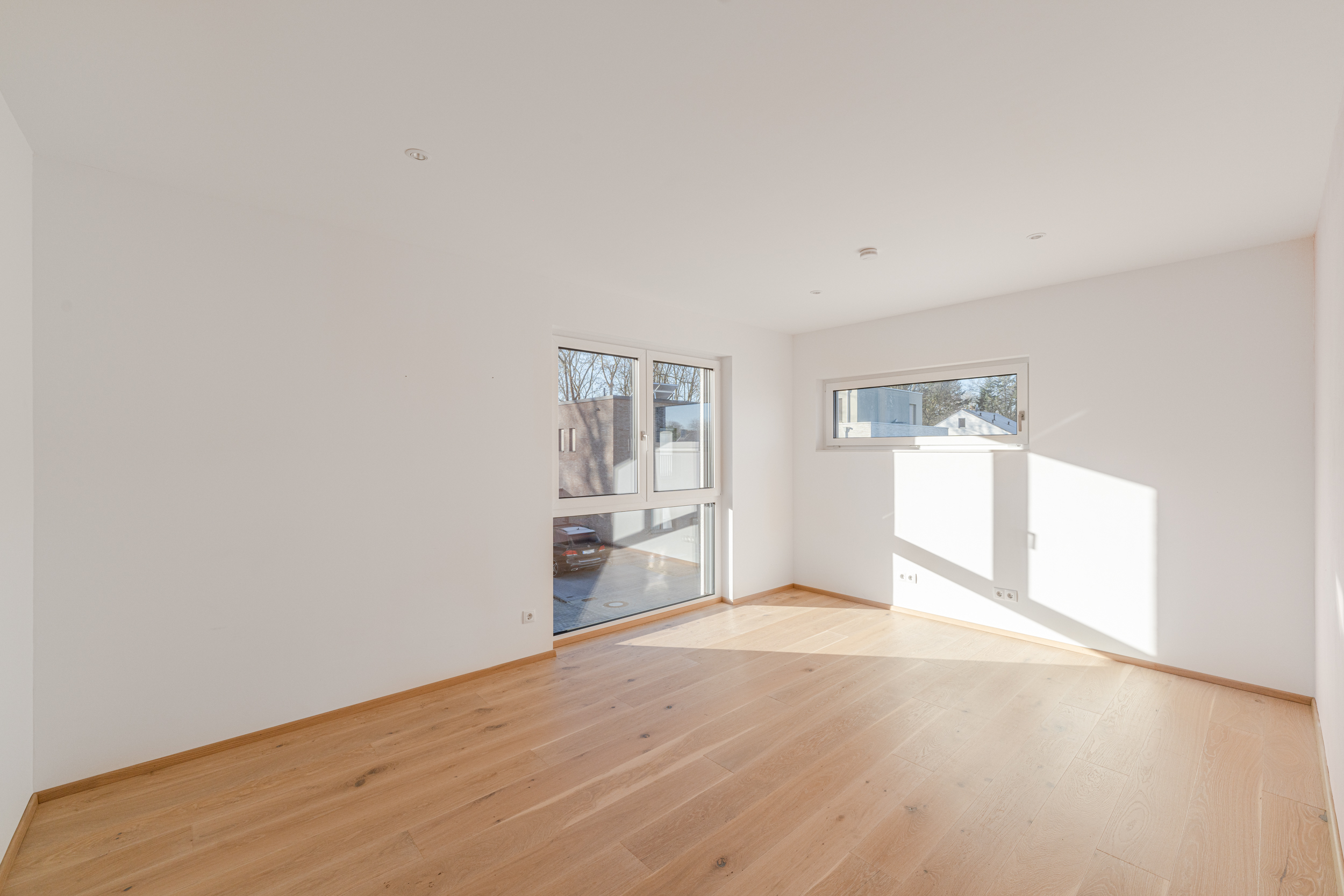 Helles, leeres Zimmer mit großen Fenstern und hellem Holzboden.