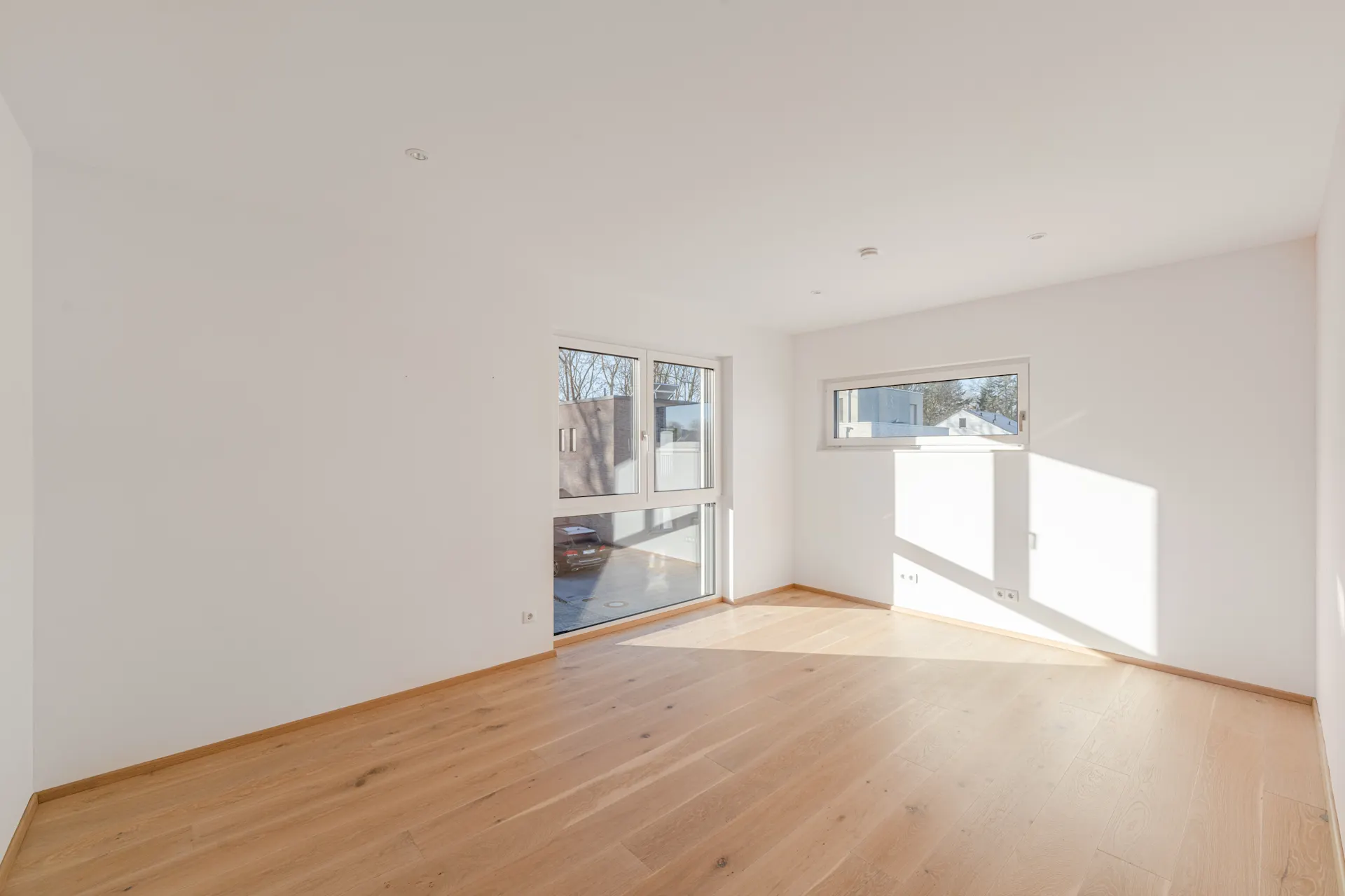 Helles, leeres Zimmer mit großen Fenstern und hellem Holzboden.