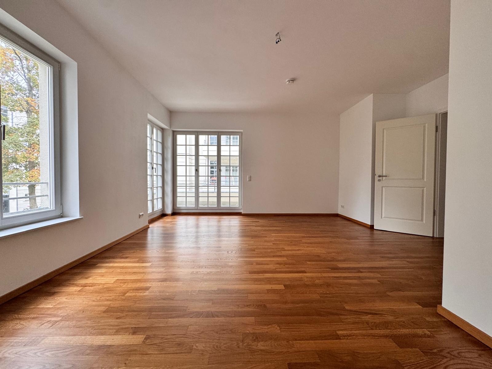 Helles, leeres Zimmer mit Holzboden, großen Fenstern und einer offenen weißen Tür.