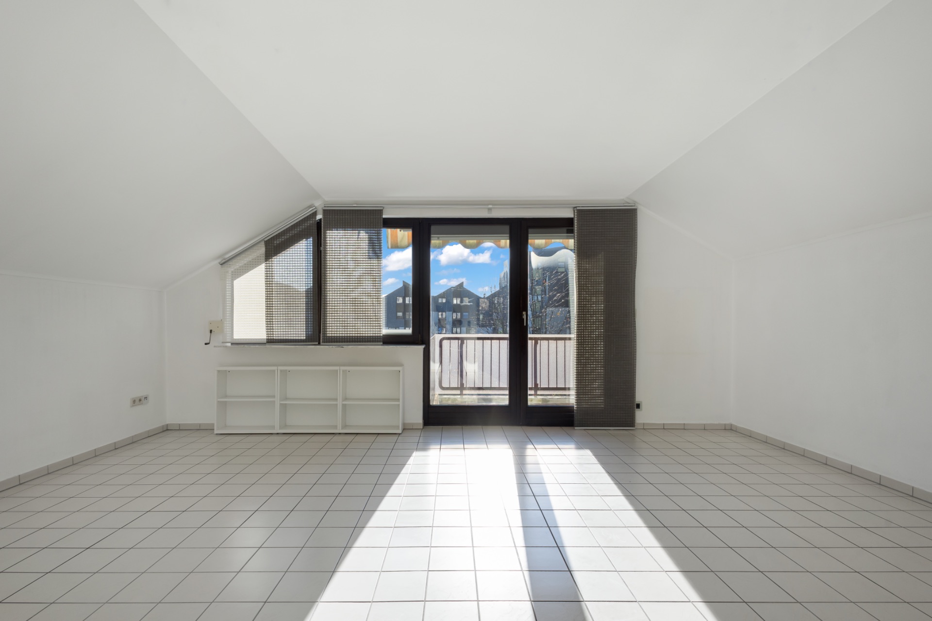 Heller Raum mit weißen Fliesen, Fensterfront und Zugang zu einem Balkon.
