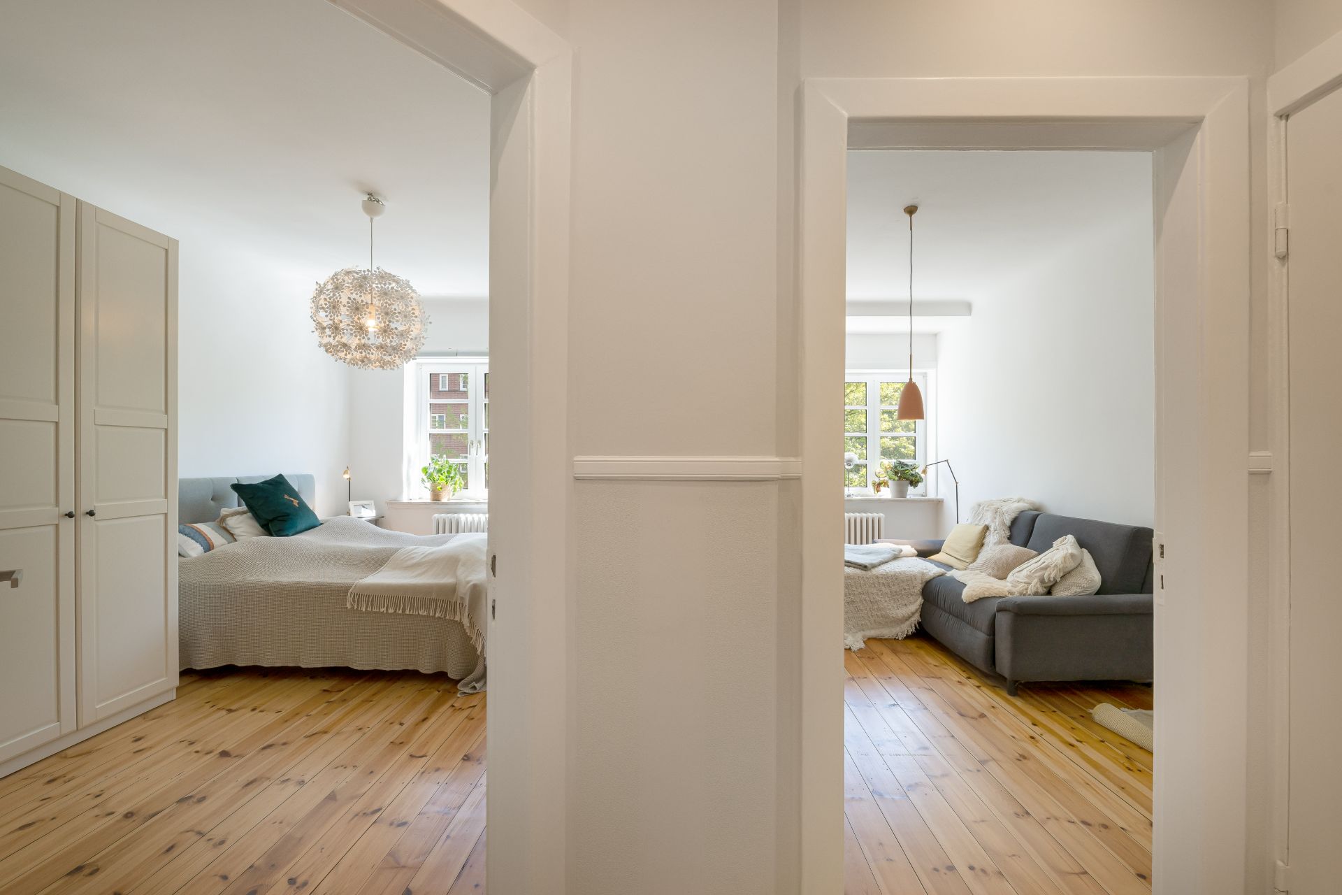 Blick durch Flur auf Schlafzimmer mit Bett links und Wohnzimmer mit Sofa rechts, Holzboden, helle Räume.