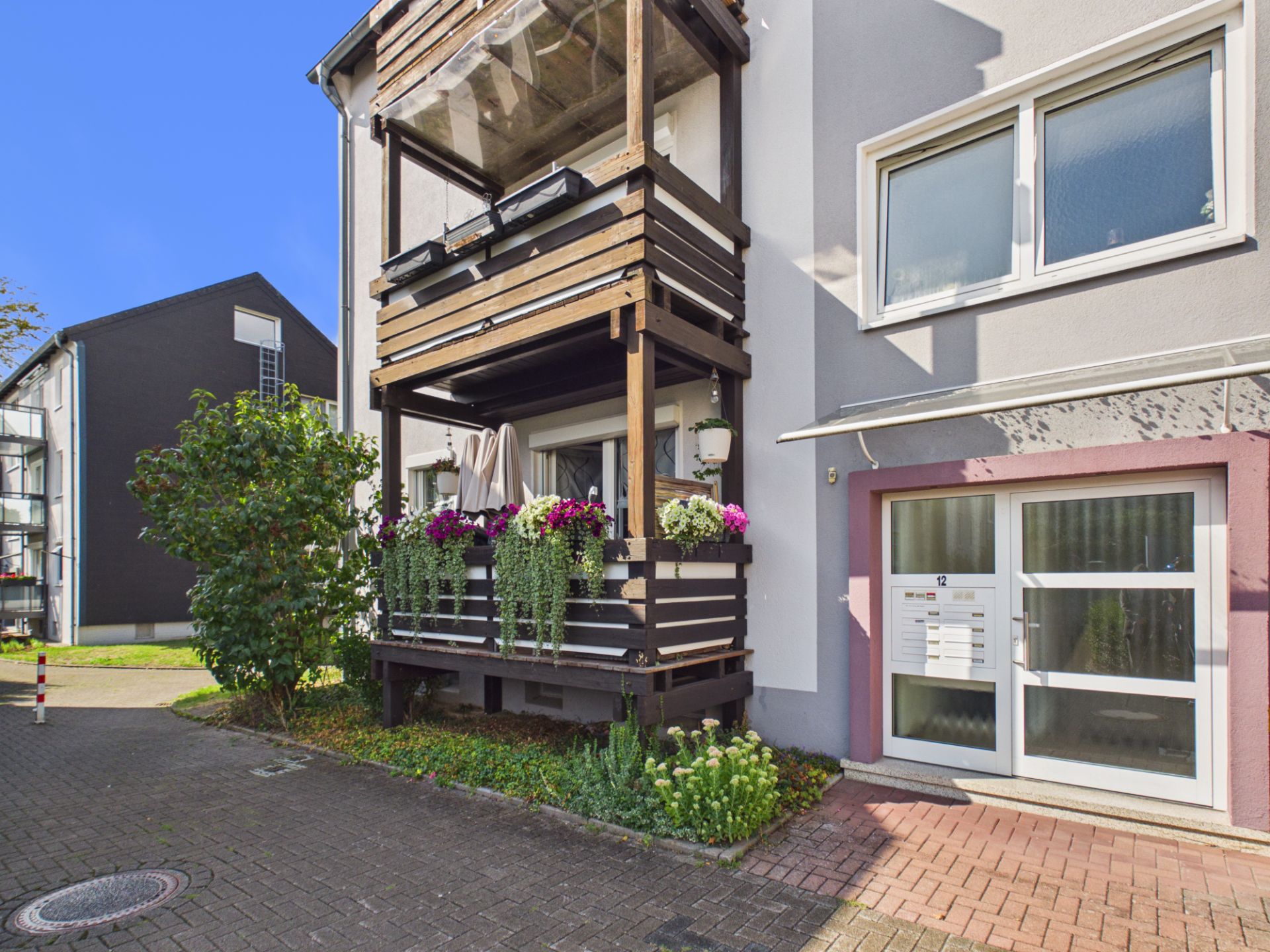 Zweistöckiger Balkon mit Blumen und Pflanzen an einem Wohnhaus, daneben Fenster und Hauseingang.