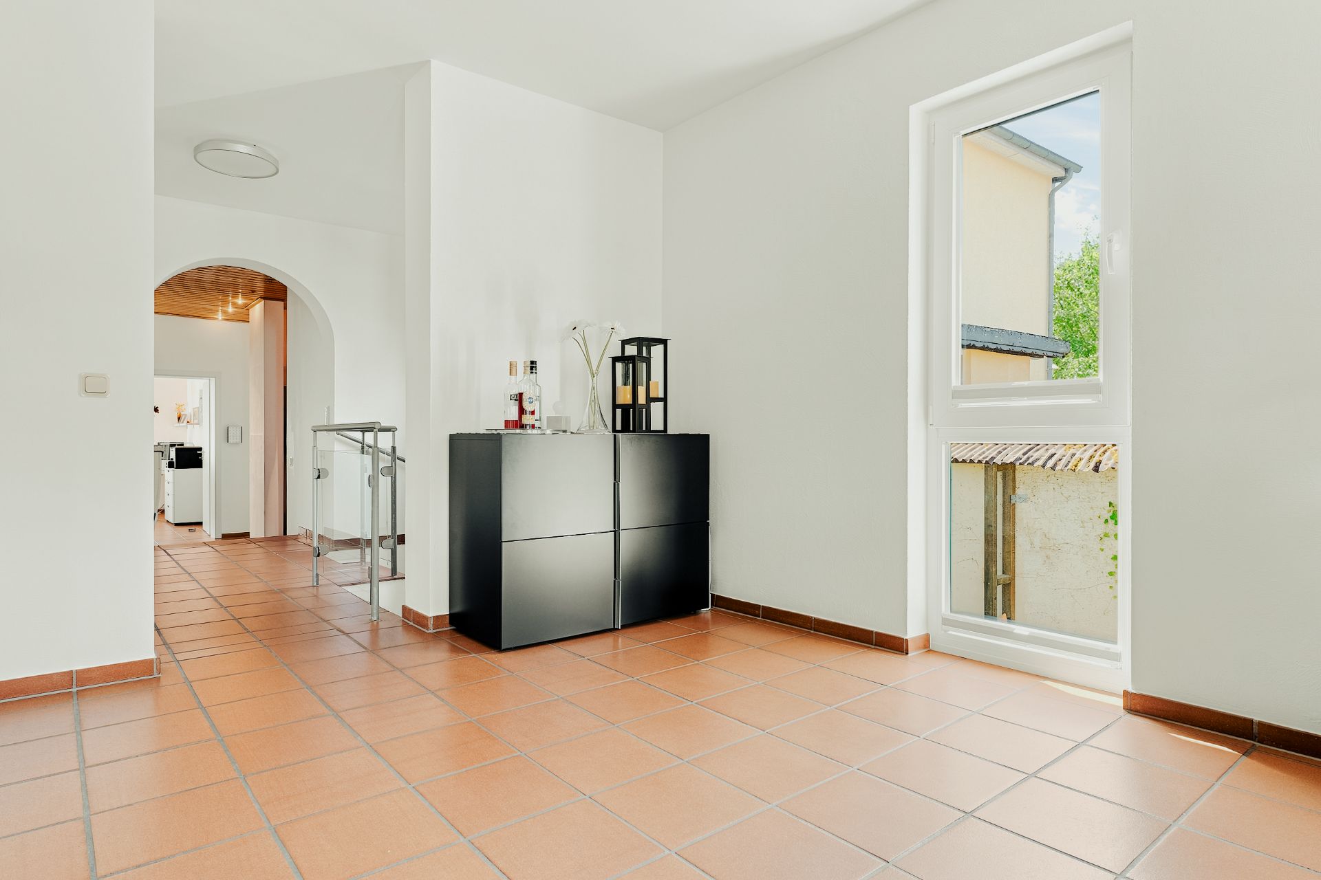 Heller Raum mit Fliesenboden, Fenster, schwarzem Sideboard und Zugang zu weiterem Zimmer.