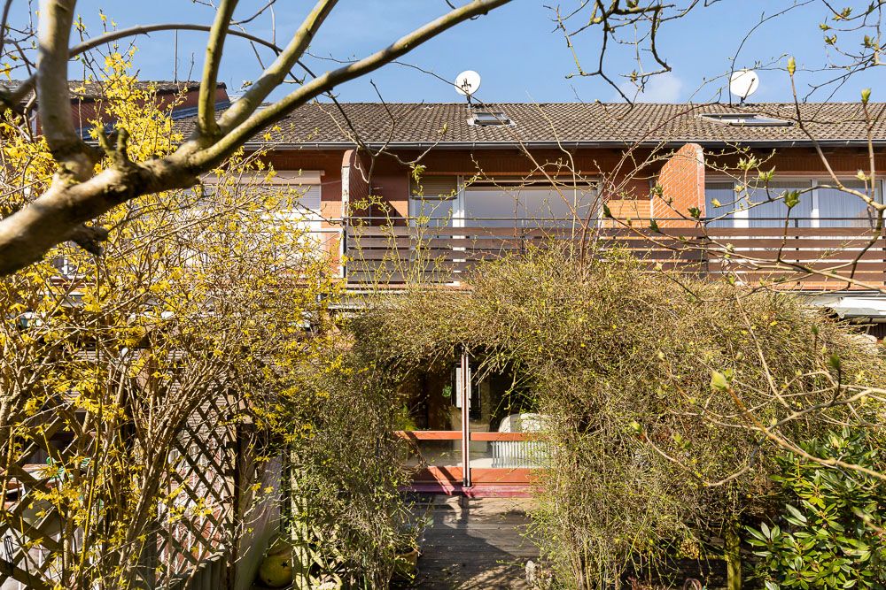 Zweistöckiges Wohnhaus mit Balkon, umgeben von Sträuchern und kahlen Ästen im Vordergrund.