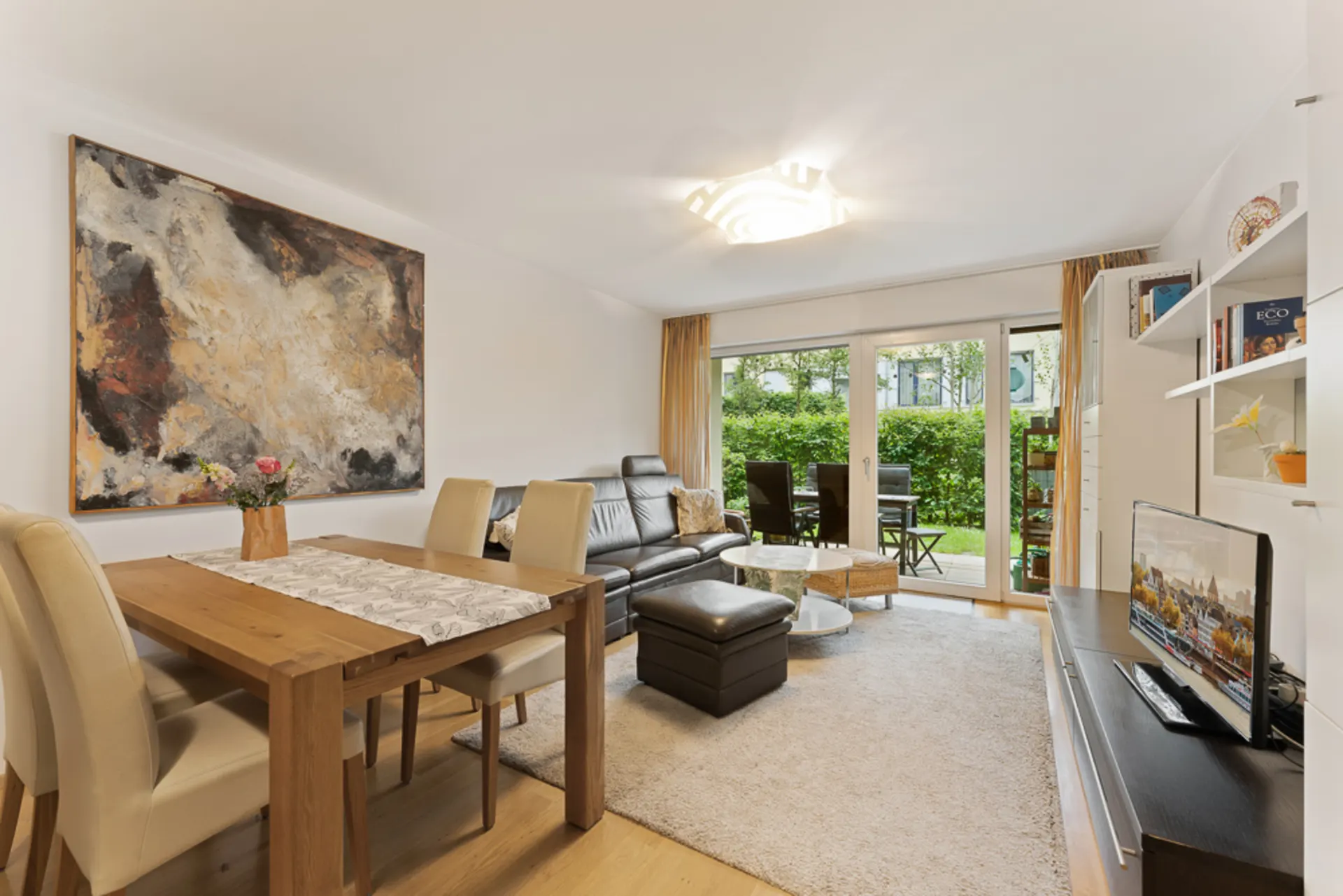 Modernes Wohnzimmer mit Esstisch, Sofa, TV, großem Gemälde und Zugang zur Terrasse mit Gartenblick.