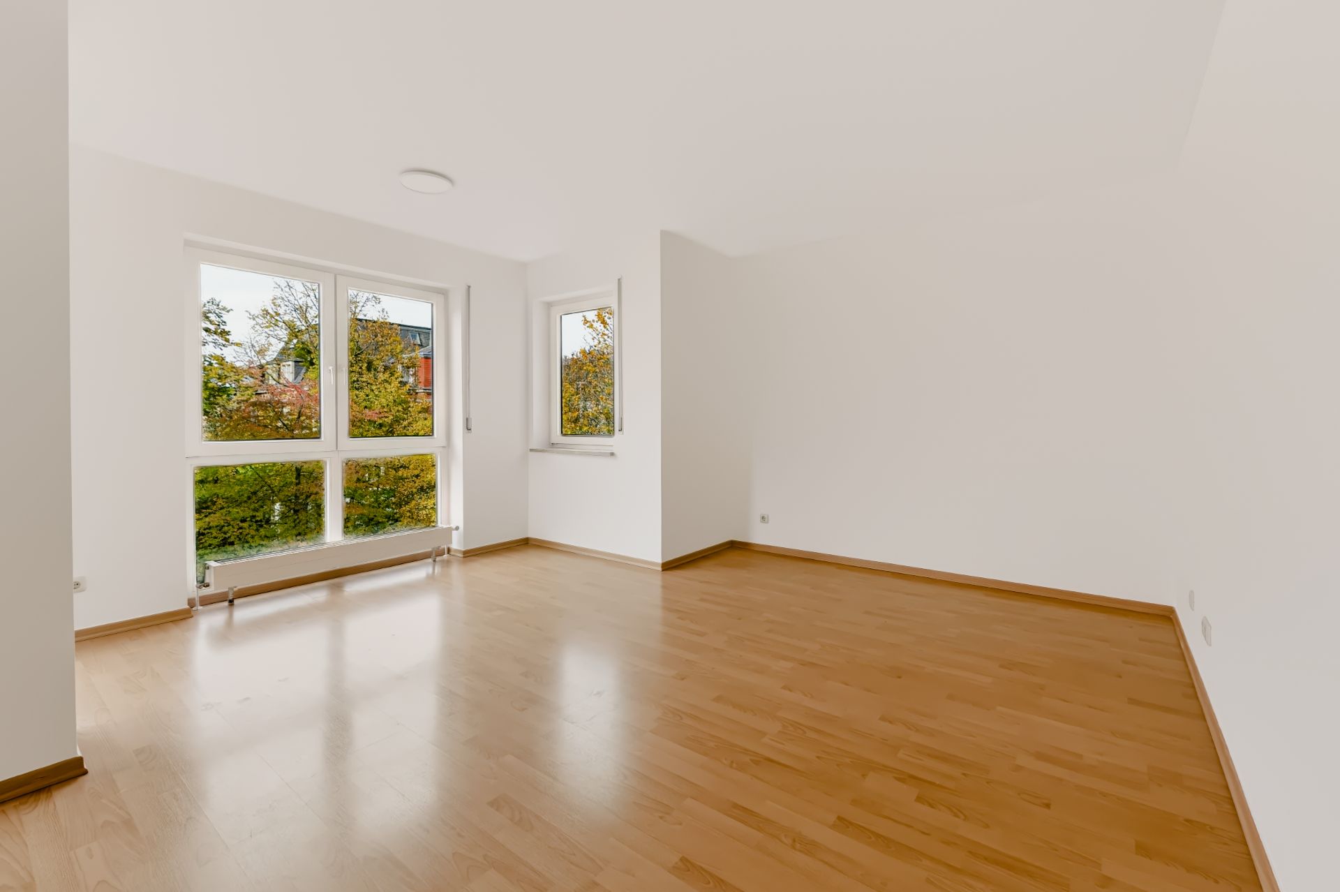 Leerer, heller Raum mit Holzfußboden und großen Fenstern mit Blick ins Grüne.