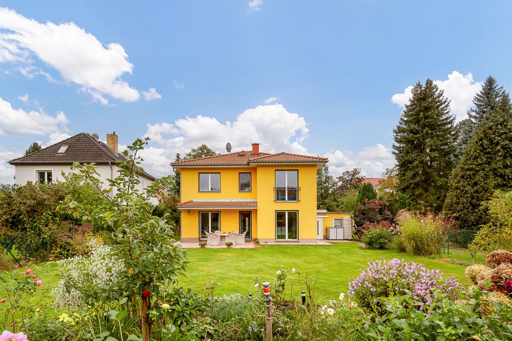 Gelbes zweistöckiges Haus mit Terrasse und großem Garten, umgeben von Bäumen und Blumen.