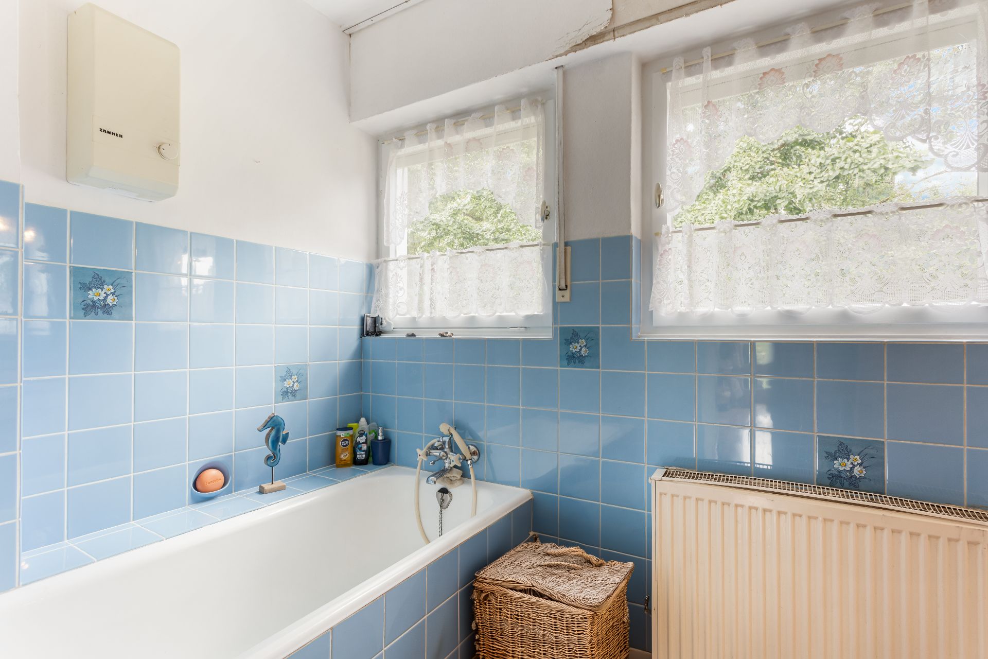 Badezimmer mit blauen Fliesen, Badewanne, zwei Fenstern und einem Wäschekorb.