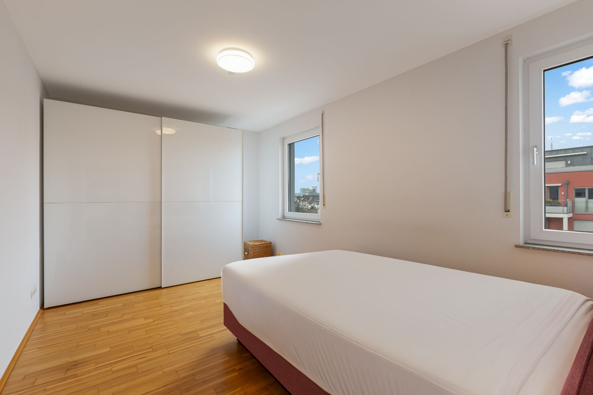 Helles Schlafzimmer mit Doppelbett, zwei Fenstern und weißem Kleiderschrank auf Holzboden.