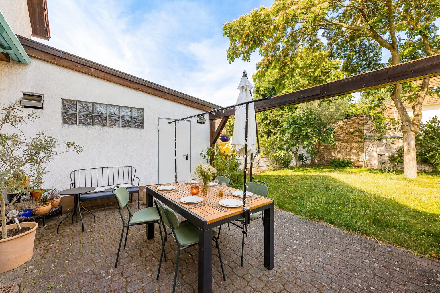 Garten mit Terrasse, Esstisch, vier Stühlen, Bank und Sonnenschirm neben Rasenfläche.