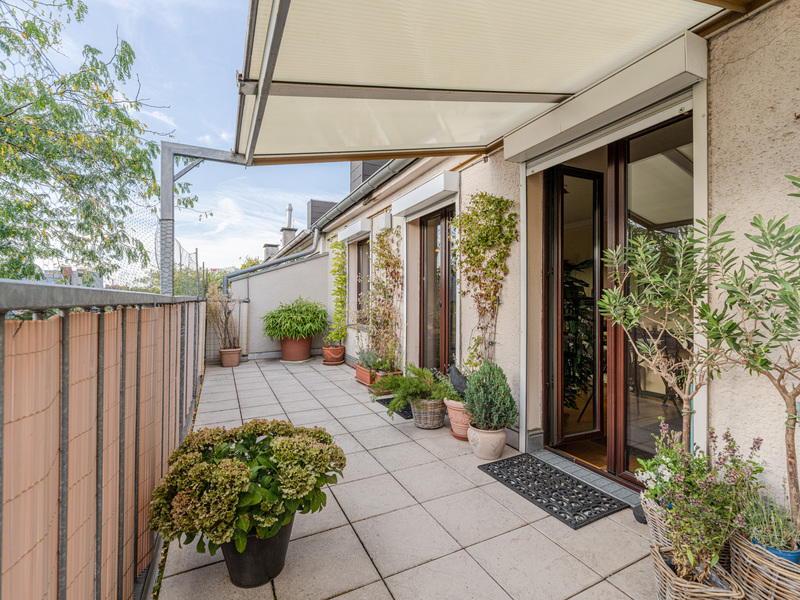 Geräumige Terrasse mit Markise, vielen Pflanzen in Töpfen und Zugang zur Wohnung durch Glastüren.