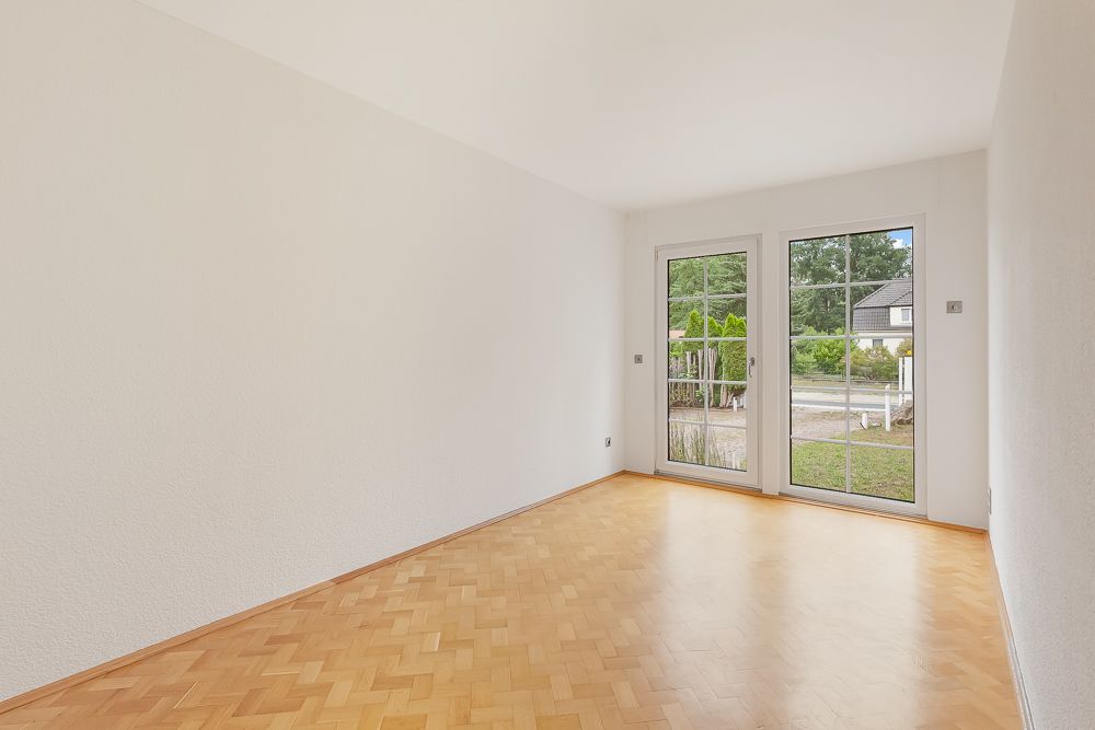 Leerer, heller Raum mit Parkettboden und großen Fenstertüren zum Garten.