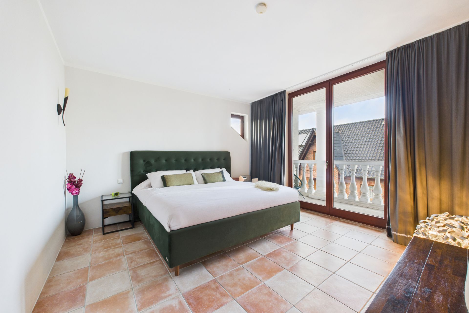 Helles Schlafzimmer mit Doppelbett, Nachttisch, Fliesenboden und großem Fenster mit Vorhängen.