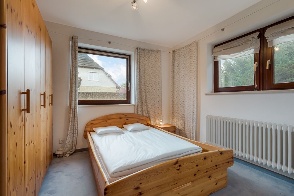Schlafzimmer mit Holzbett, Kleiderschrank, Nachttisch und großen Fenstern mit Vorhängen.