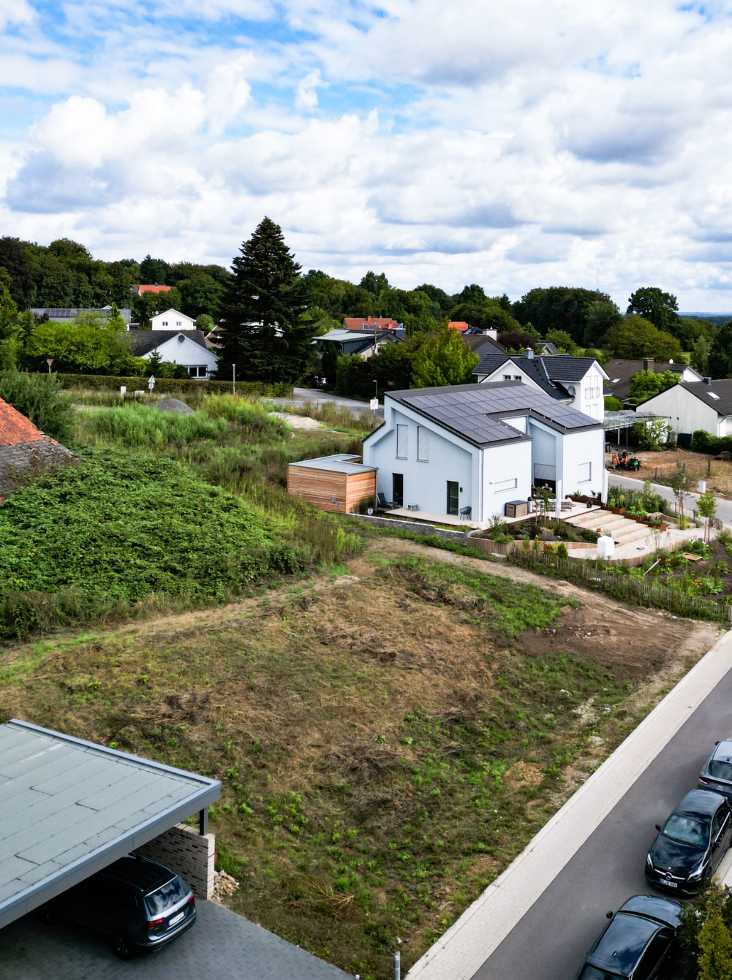 Modernes weißes Einfamilienhaus mit Garten und Solarpanels, umgeben von Grünflächen und Nachbarhäusern.