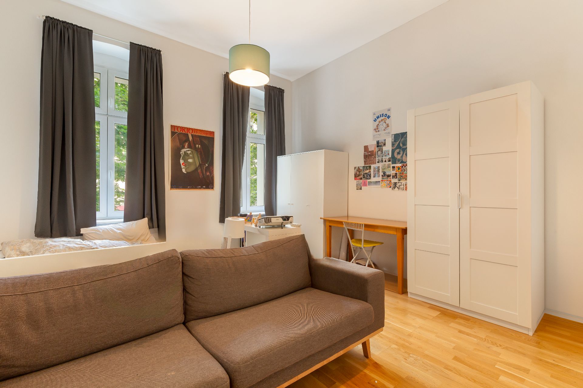 Helles Zimmer mit braunem Sofa, großem Schrank, Schreibtisch und zwei Fenstern mit dunklen Vorhängen.