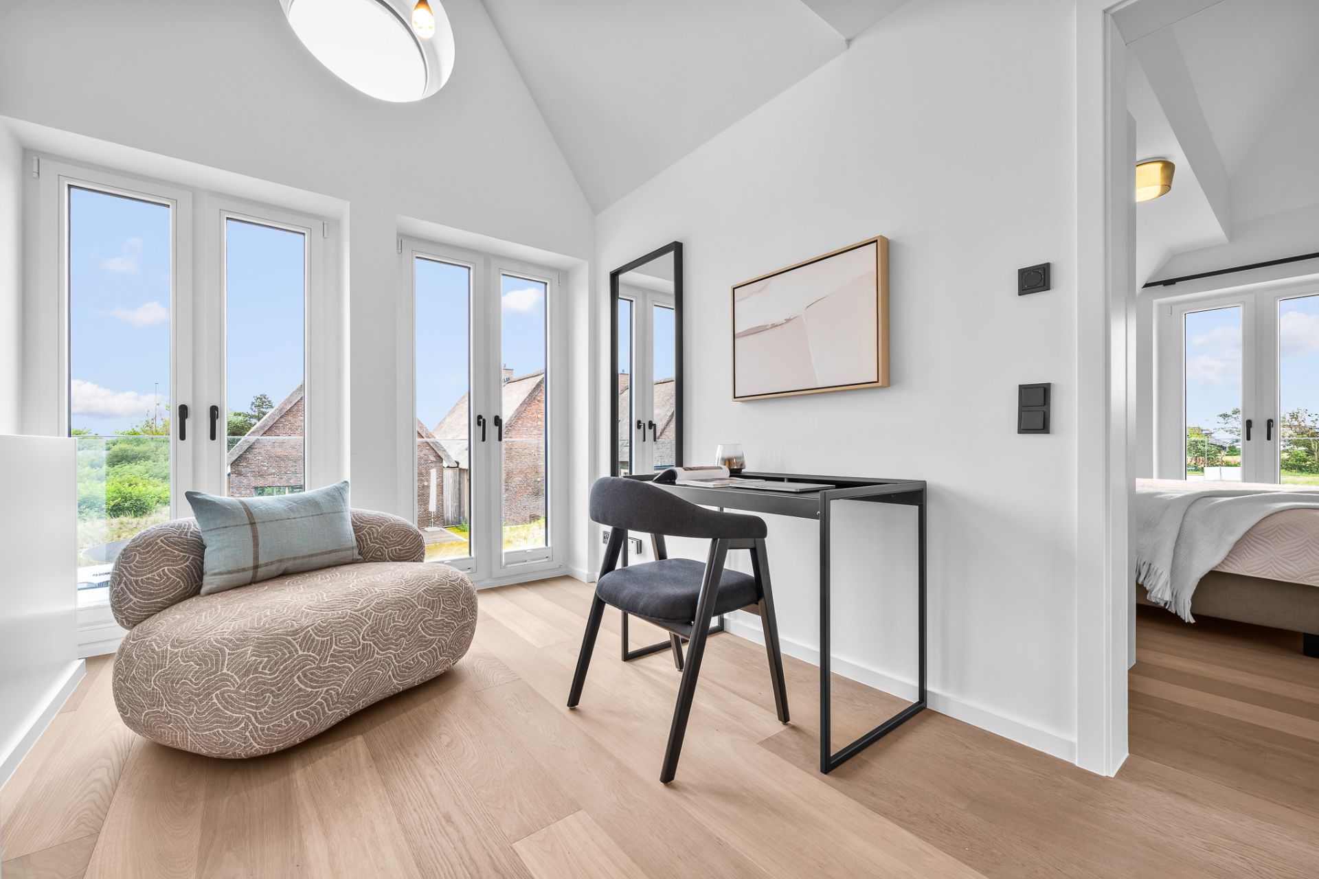 Helles Zimmer mit Sessel, Schreibtisch, Stuhl und großen Fenstern, daneben ein Schlafzimmer.