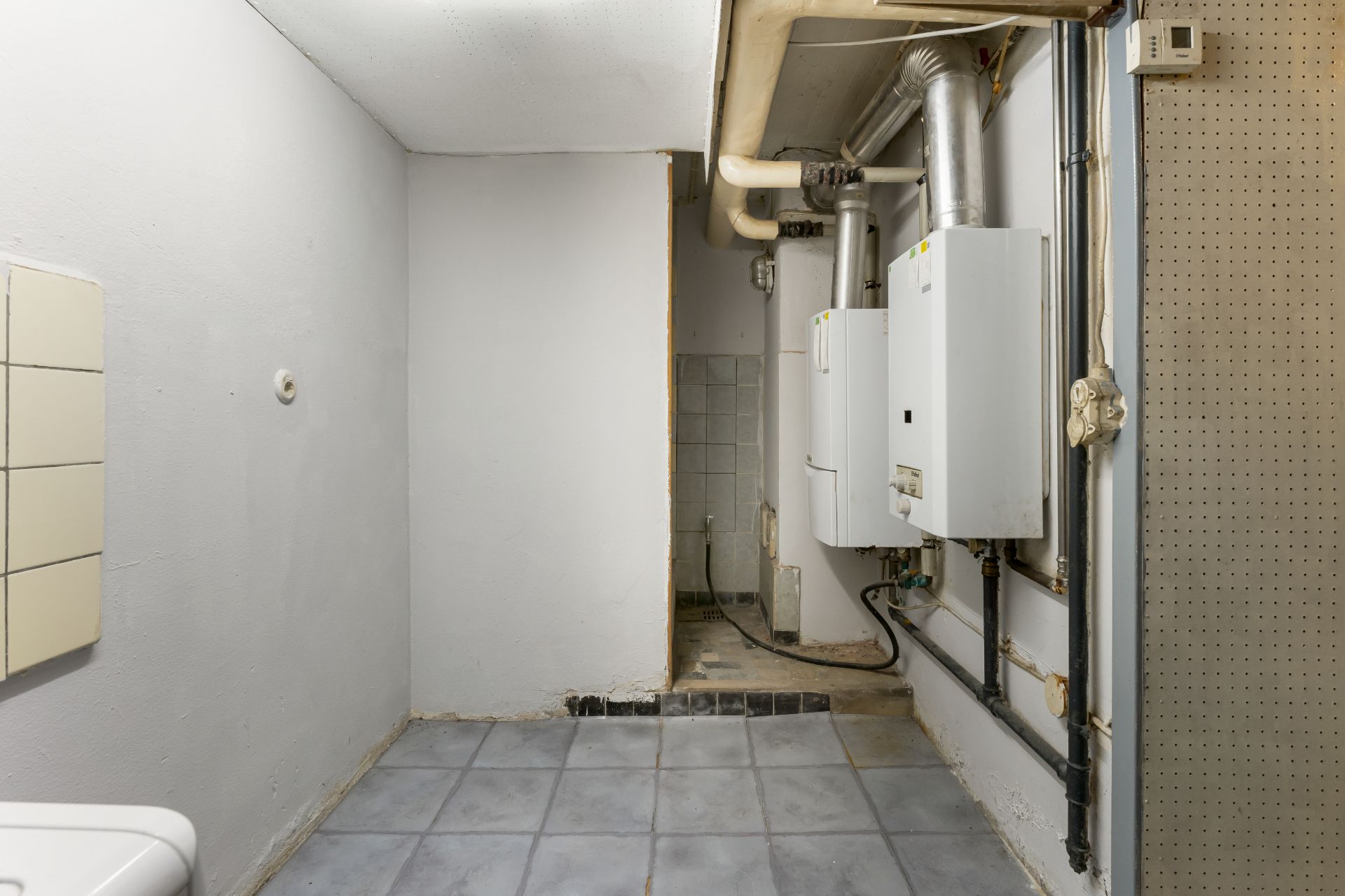 Kellerraum mit Fliesenboden, Heizungsanlage und Rohrleitungen an der Wand.
