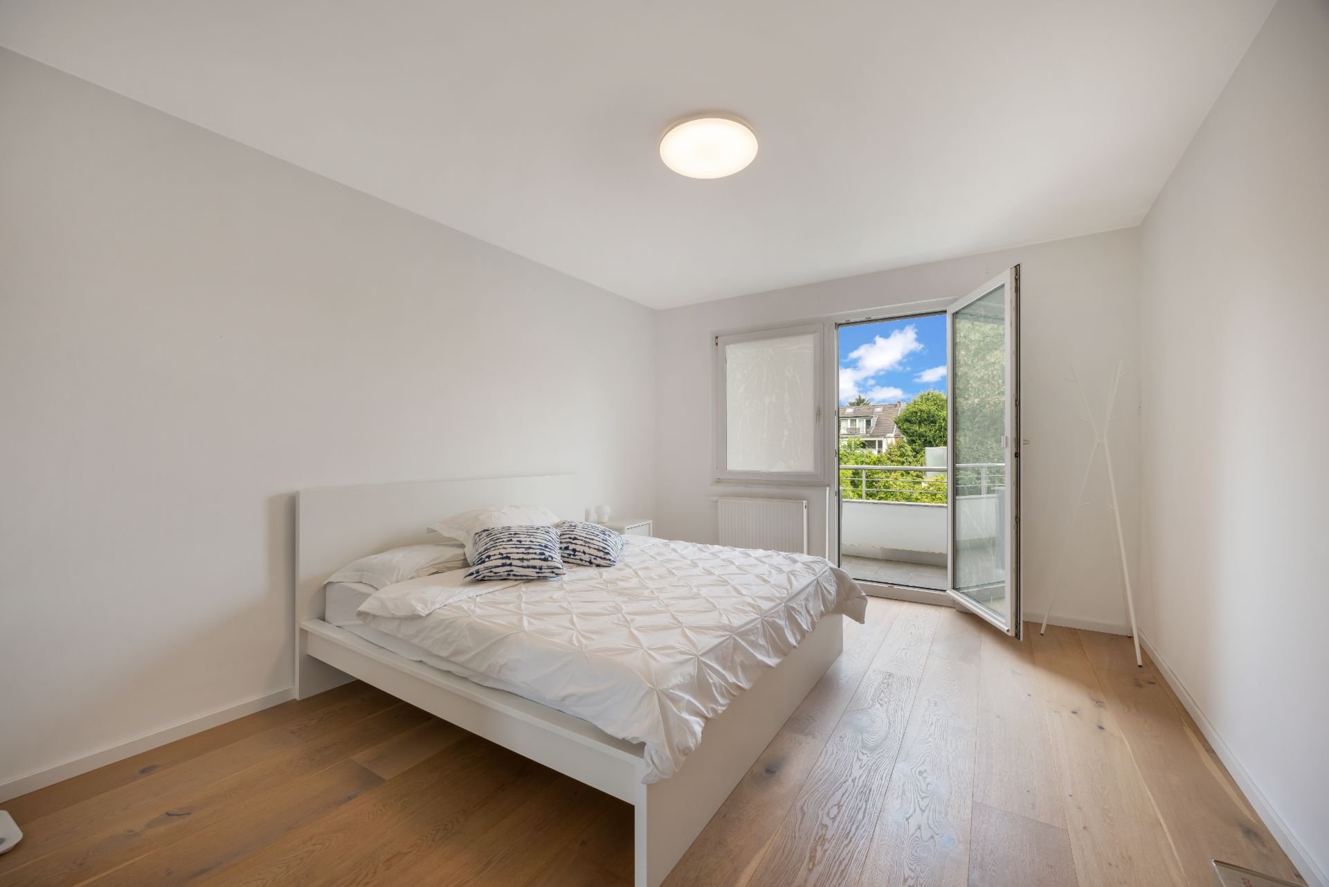 Helles Schlafzimmer mit Doppelbett, Holzboden und Zugang zu einem Balkon mit Ausblick ins Grüne.