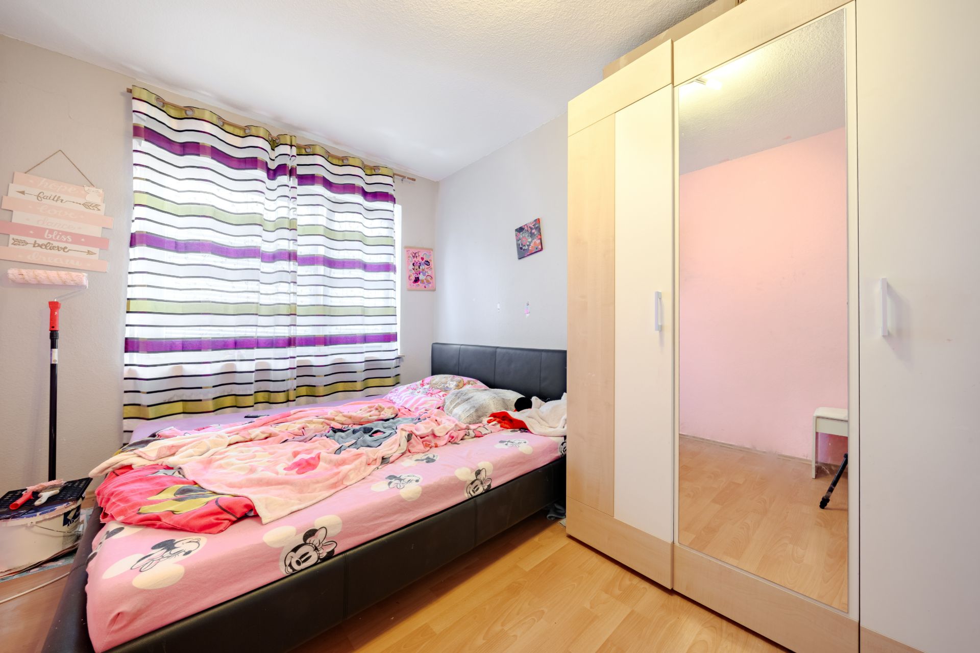 Helles Schlafzimmer mit Bett, bunter Bettwäsche, Kleiderschrank mit Spiegel und Fenster mit gestreiften Vorhängen.