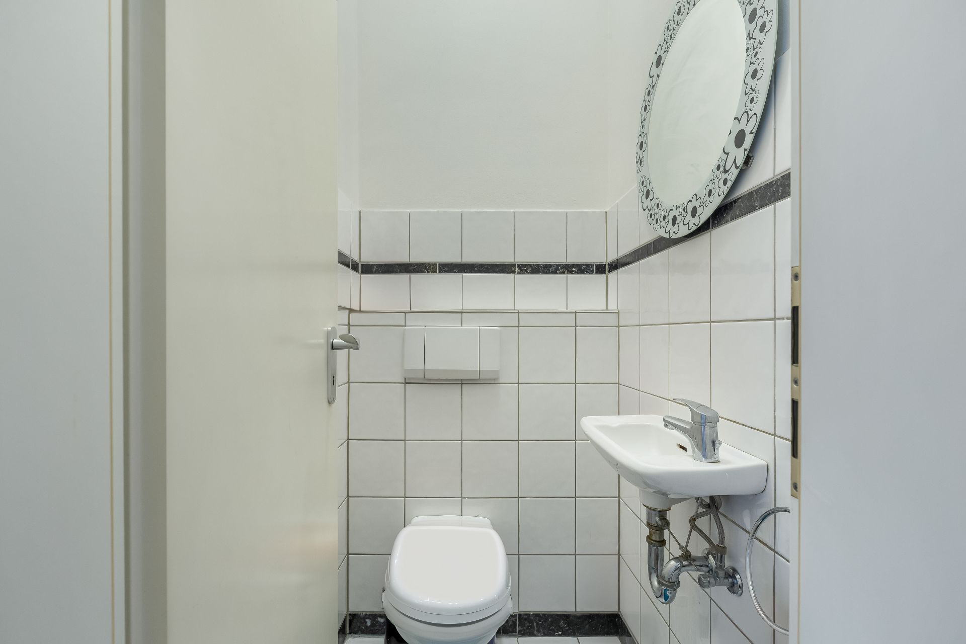 Kleines, gefliestes WC mit weißer Toilette, kleinem Waschbecken und rundem Spiegel.