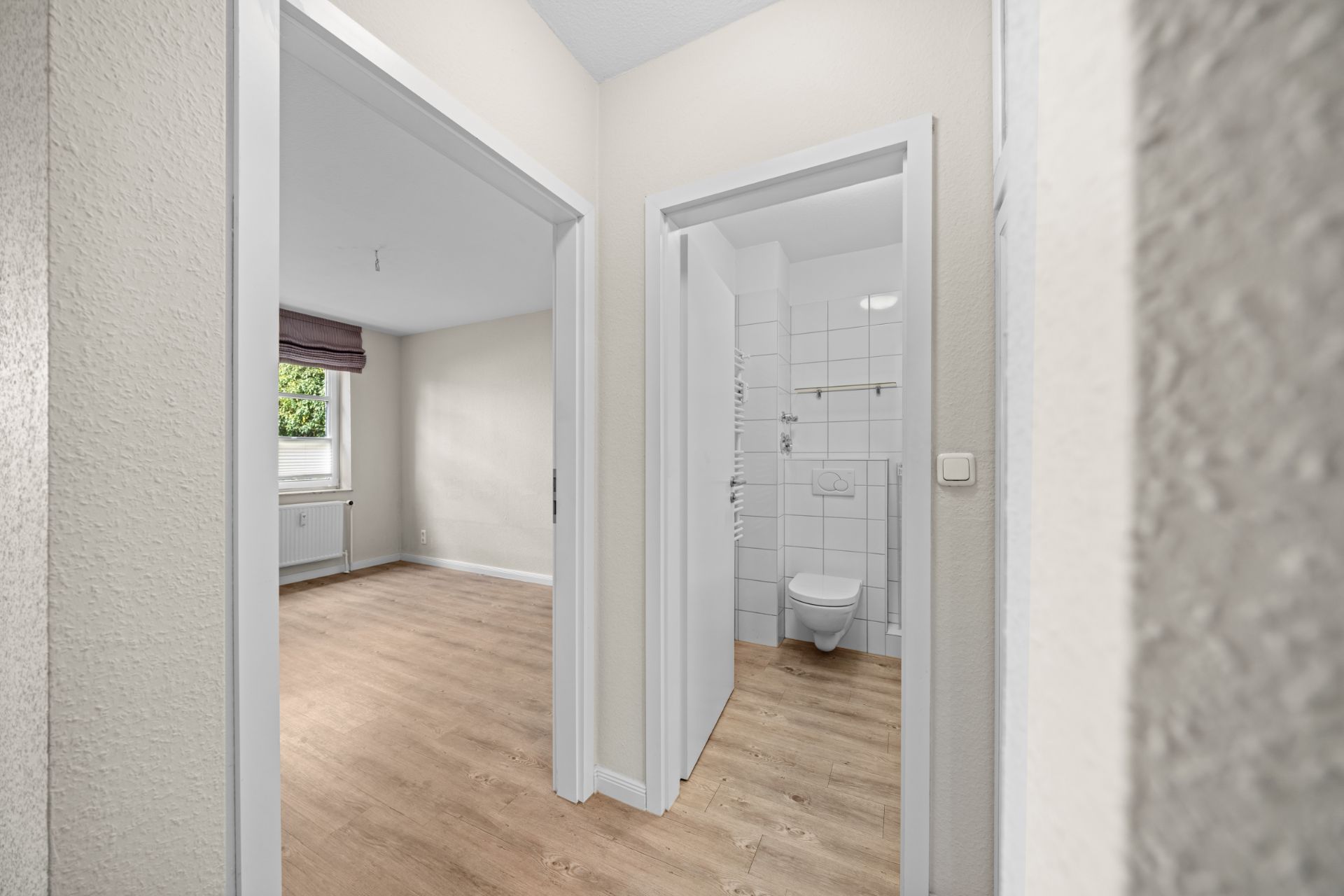 Flur mit Blick auf ein leeres Zimmer und ein weiß gefliestes Bad mit Toilette.