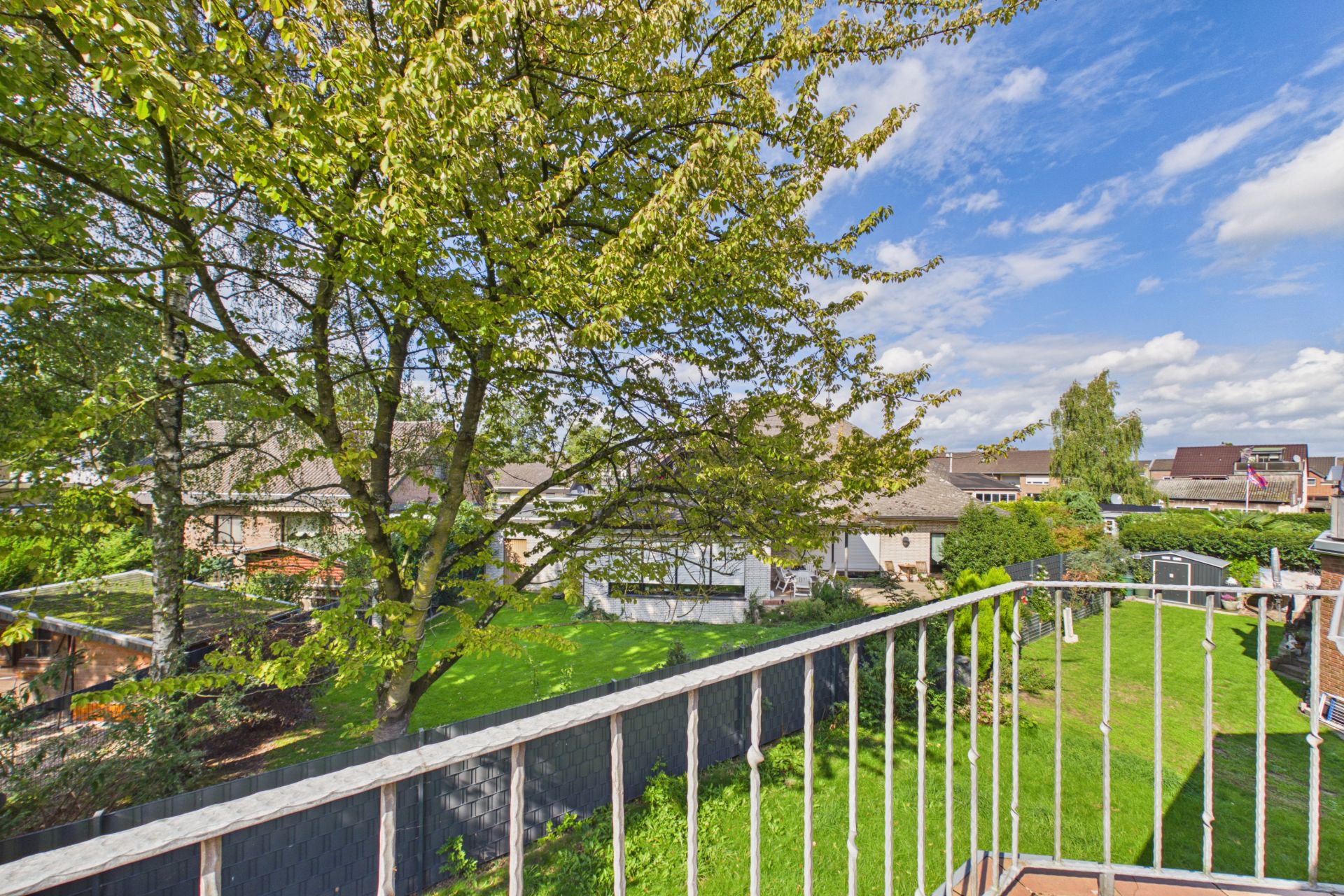 Blick von einem Balkon auf einen grünen Garten mit Bäumen und benachbarten Häusern.