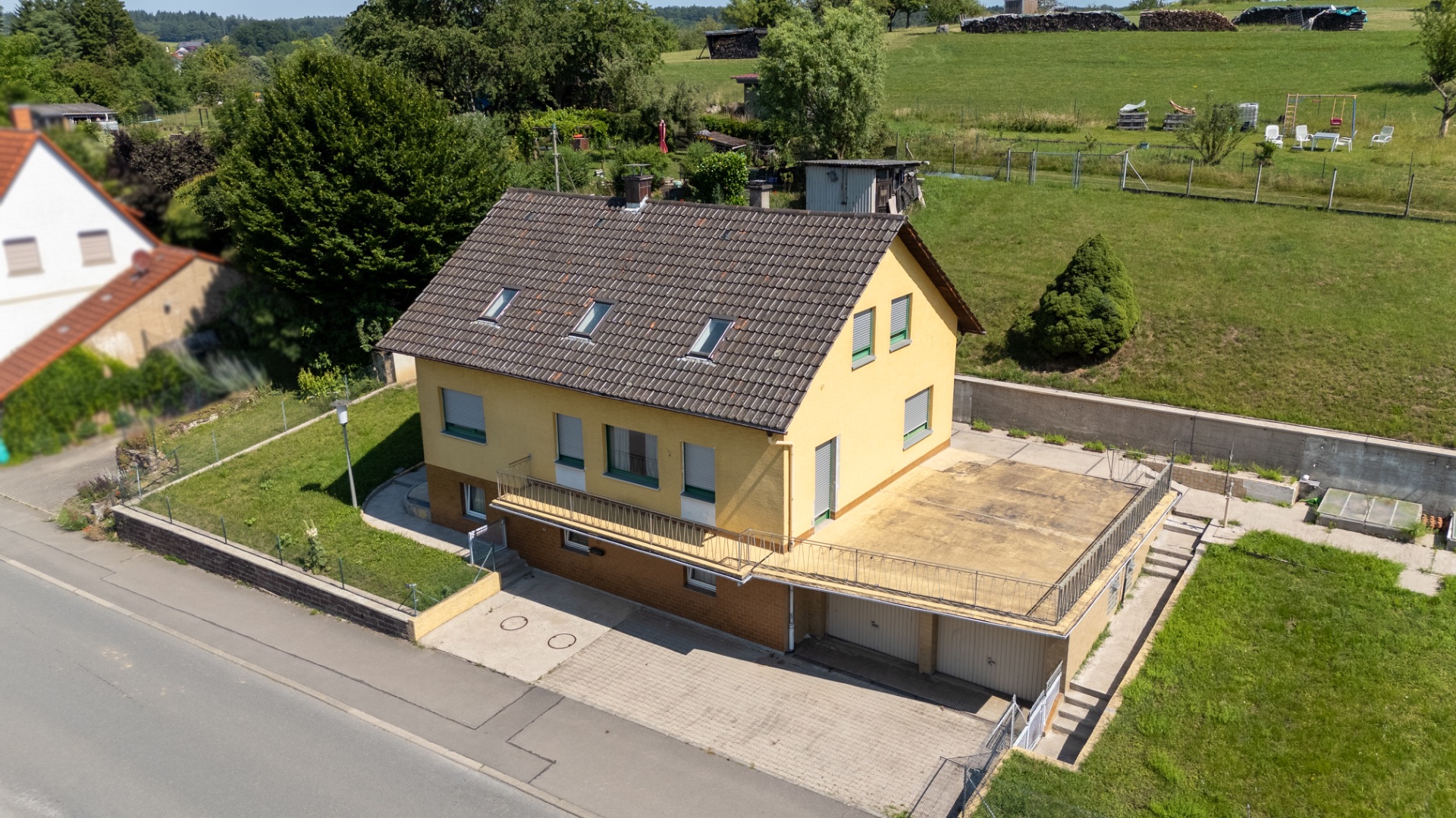 Gelbes Wohnhaus mit Schrägdach, Garage und großem gepflasterten Vorplatz, umgeben von Garten.