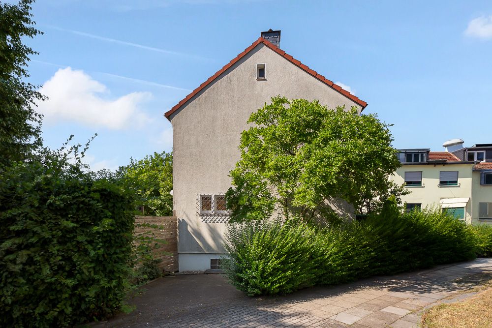 Seitenansicht eines Wohnhauses mit Satteldach, umliegenden Büschen und Bäumen bei sonnigem Wetter.