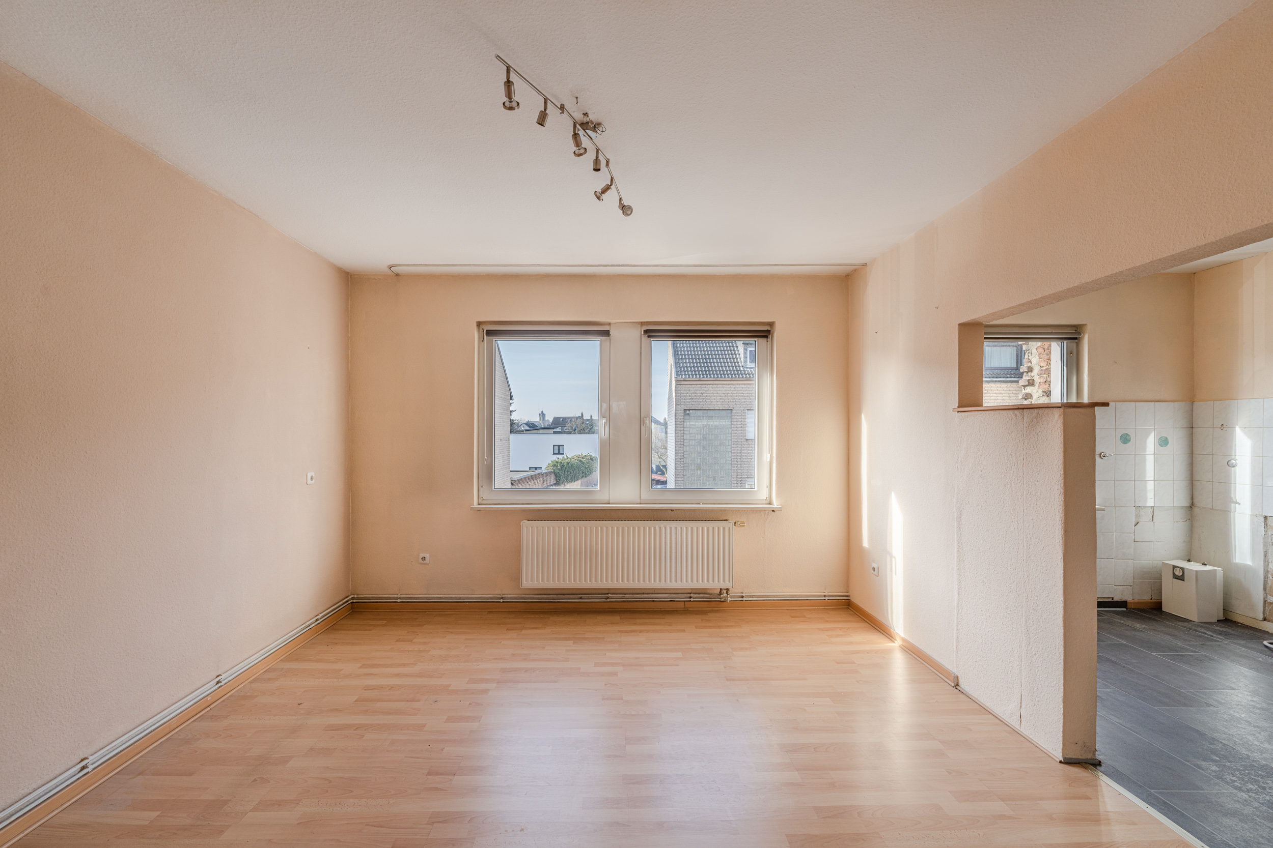 Leeres Wohnzimmer mit hellen Wänden, Laminatboden und zwei Fenstern, rechts offener Zugang zur Küche.