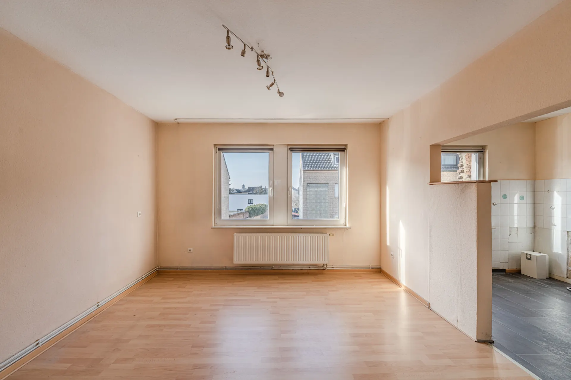 Leeres Wohnzimmer mit hellen Wänden, Laminatboden und zwei Fenstern, rechts offener Zugang zur Küche.