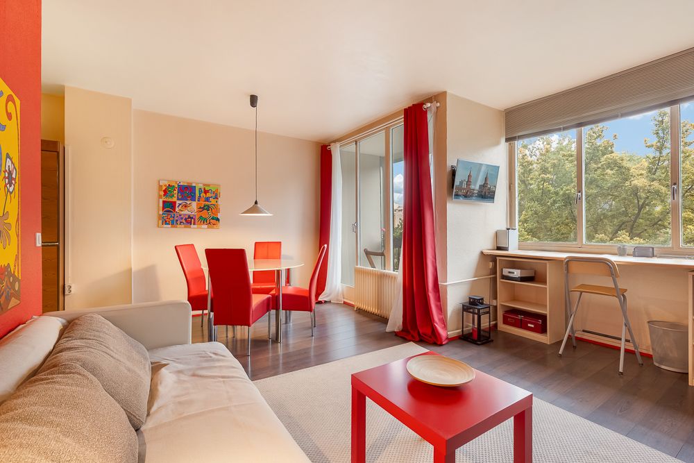 Modernes Wohnzimmer mit rotem Tisch, Sofa, Esstisch mit roten Stühlen und großem Fenster mit Blick ins Grüne.