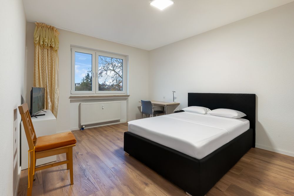 Helles Schlafzimmer mit Doppelbett, Schreibtisch, Stuhl, Fernseher und Fenster mit Vorhang.