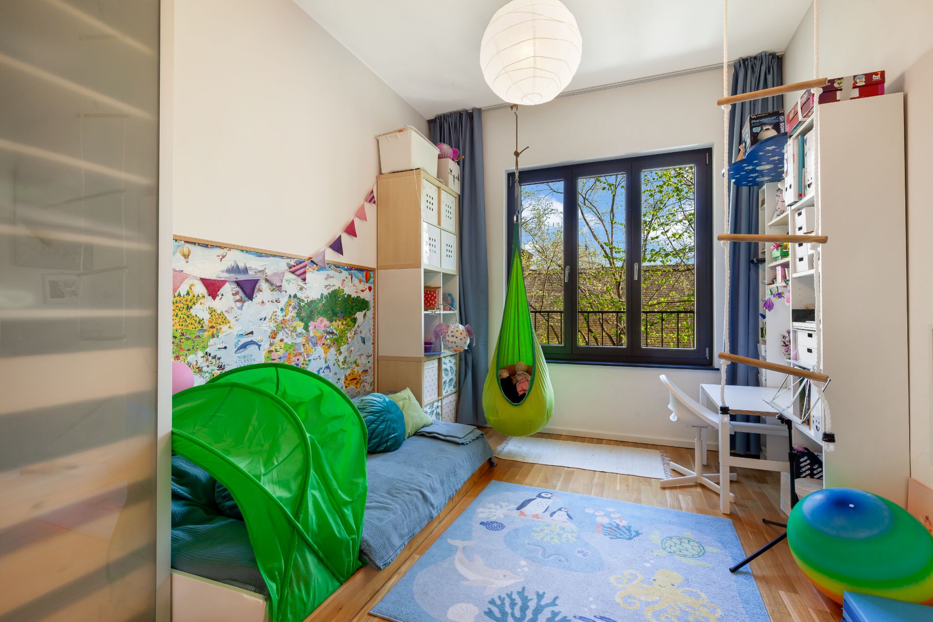 Helles Kinderzimmer mit Spielzelt, Hängesessel, Teppich und großem Fenster.