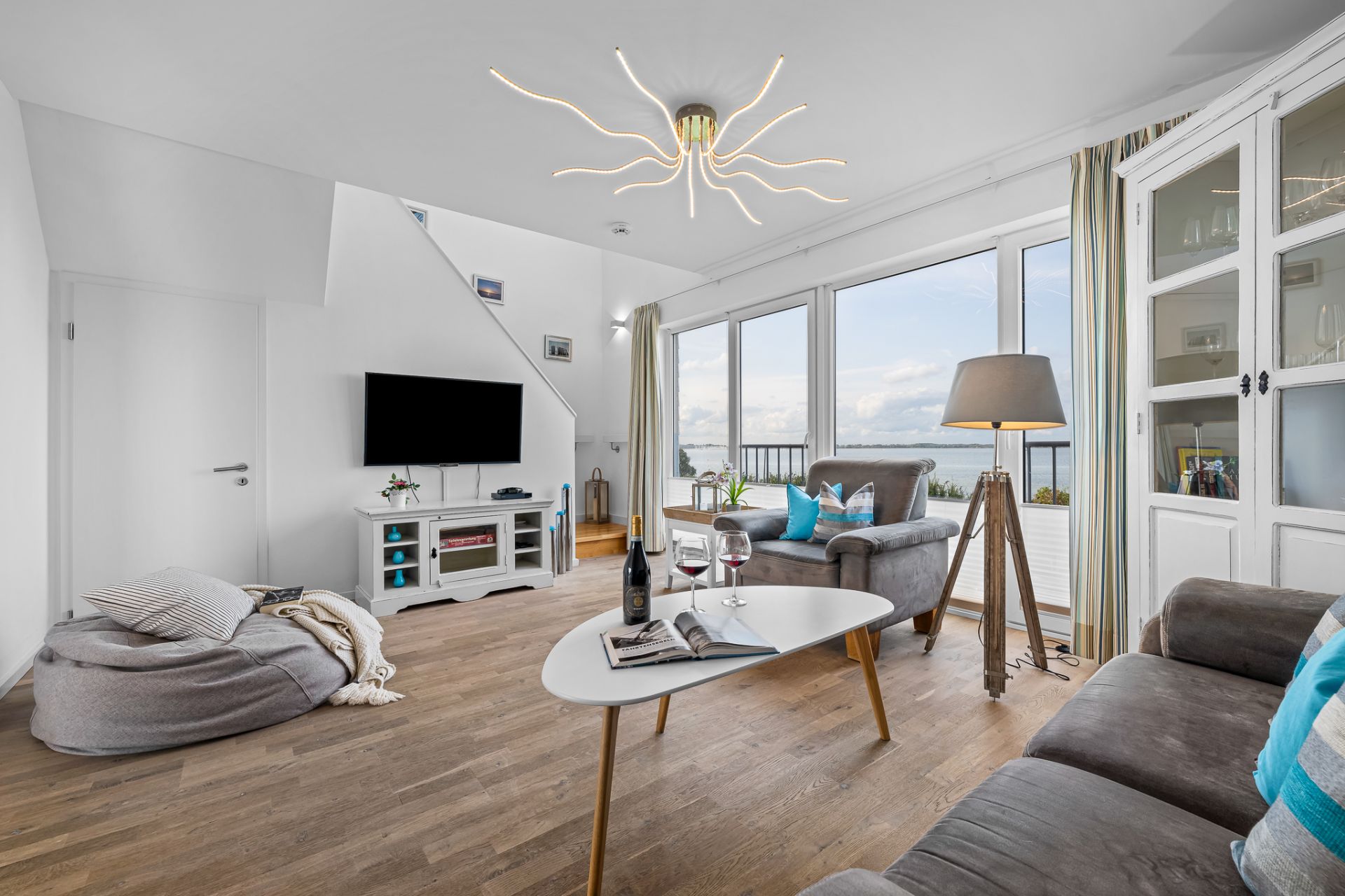 Helles Wohnzimmer mit Sofa, Sessel, Couchtisch, Stehlampe und großen Fenstern mit Aussicht.